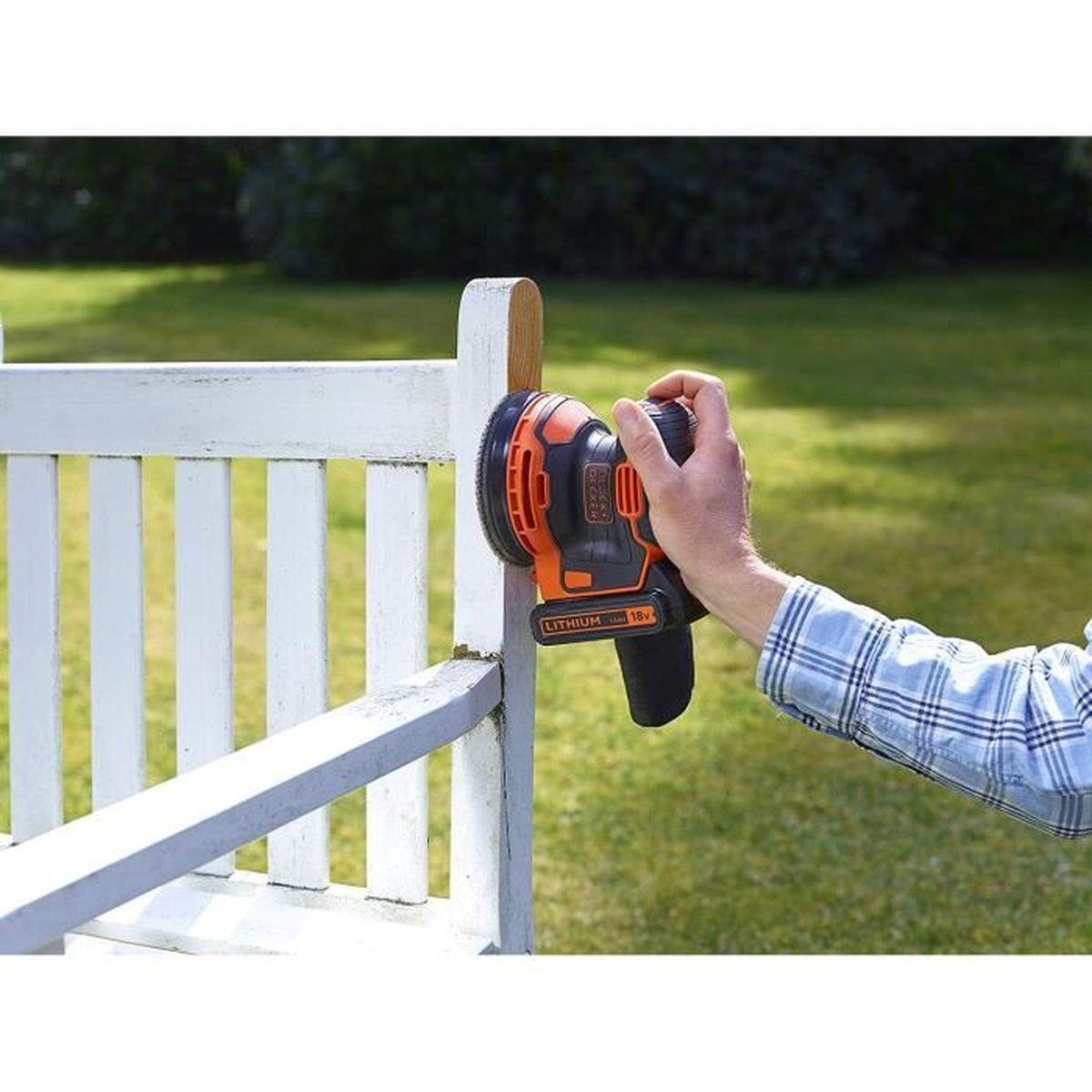 Personne ponçant un banc en bois blanc avec une ponceuse électrique noire et orange. Fond d'herbe verte.