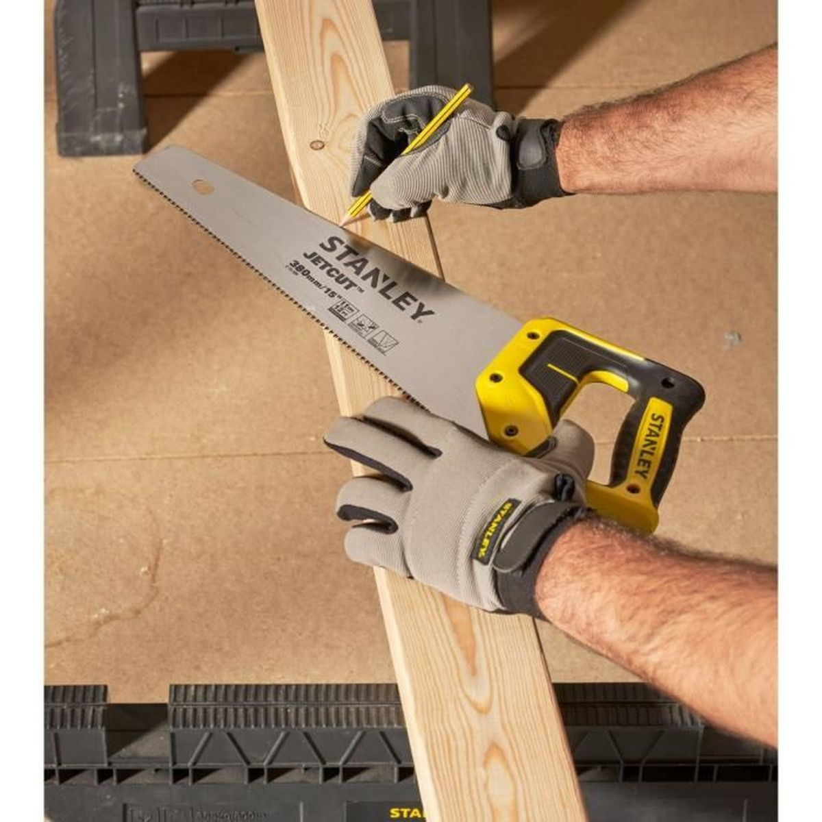 Hände mit Handschuhen, die eine Säge benutzen, um ein Holzbrett zu schneiden. Ein Bleistift markiert die Schnittlinie.