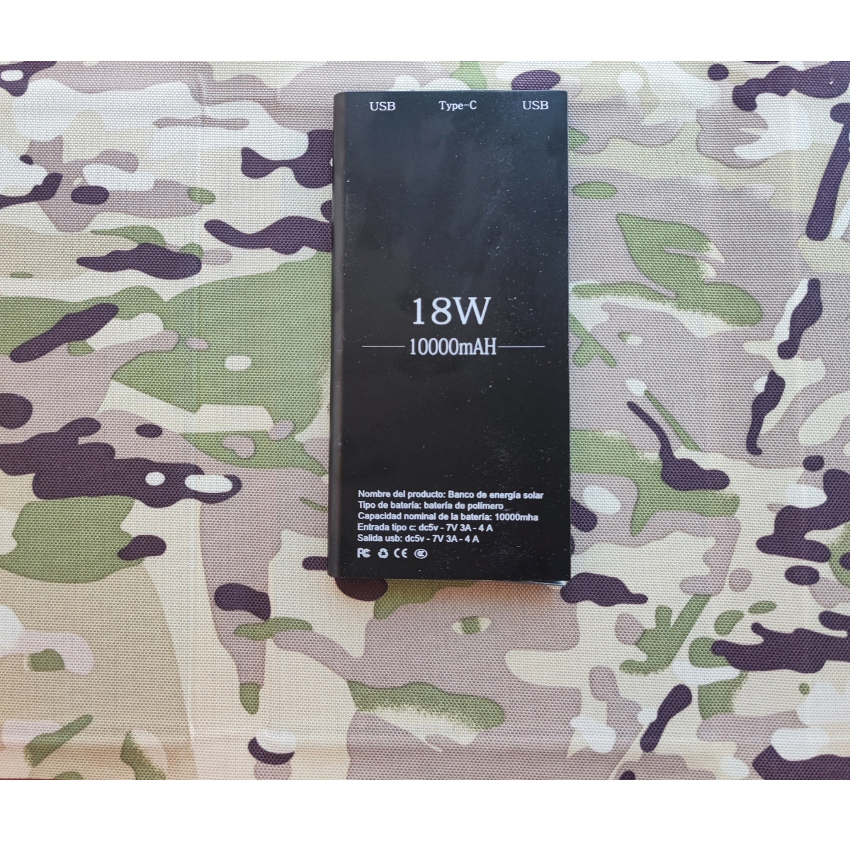 Banco de energía negro con impresión 18W-10000mAh, puertos USB/Tipo-C, sobre fondo camuflaje.