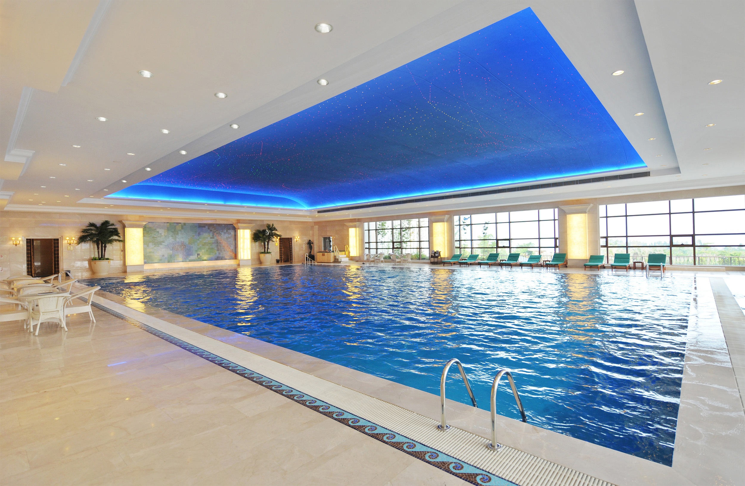 Hallenbad mit blau beleuchteter Decke. Klares Wasser, weiße Fliesen und große Fenster.
