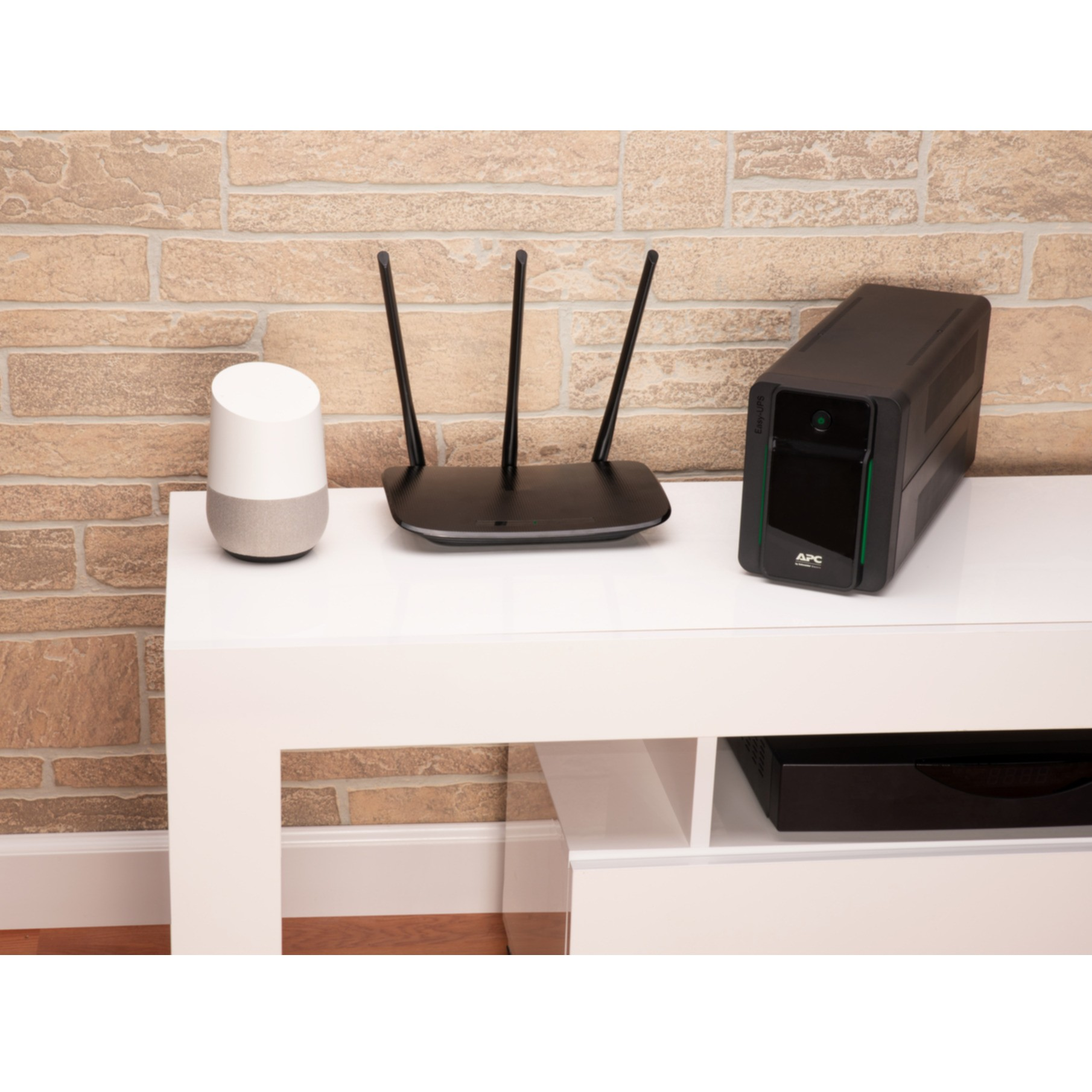Głośnik smart, router i czarny UPS na białej półce, na tle ceglanej ściany.