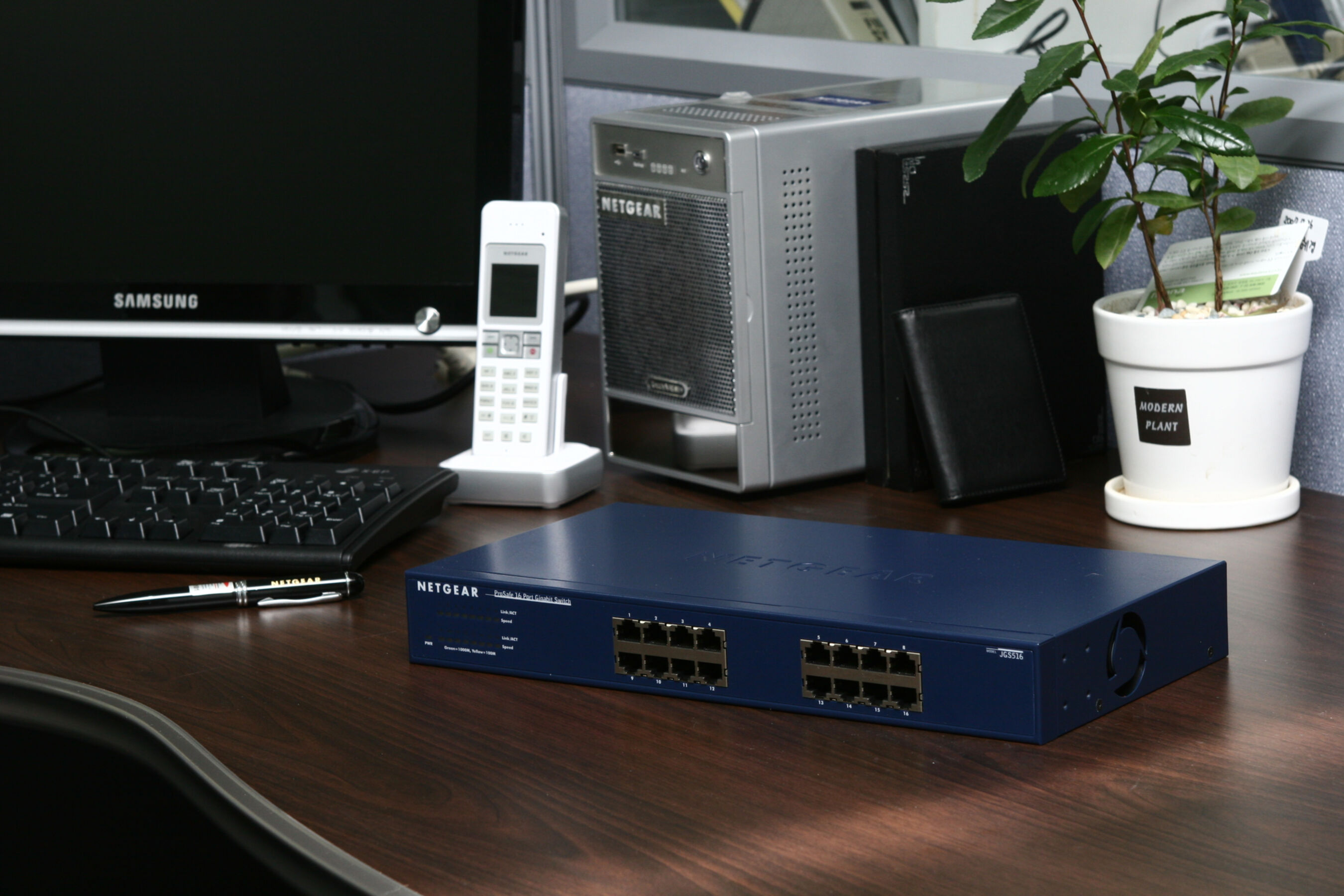 Scrivania con computer, telefono e switch di rete. Una pianta è posta sulla scrivania.