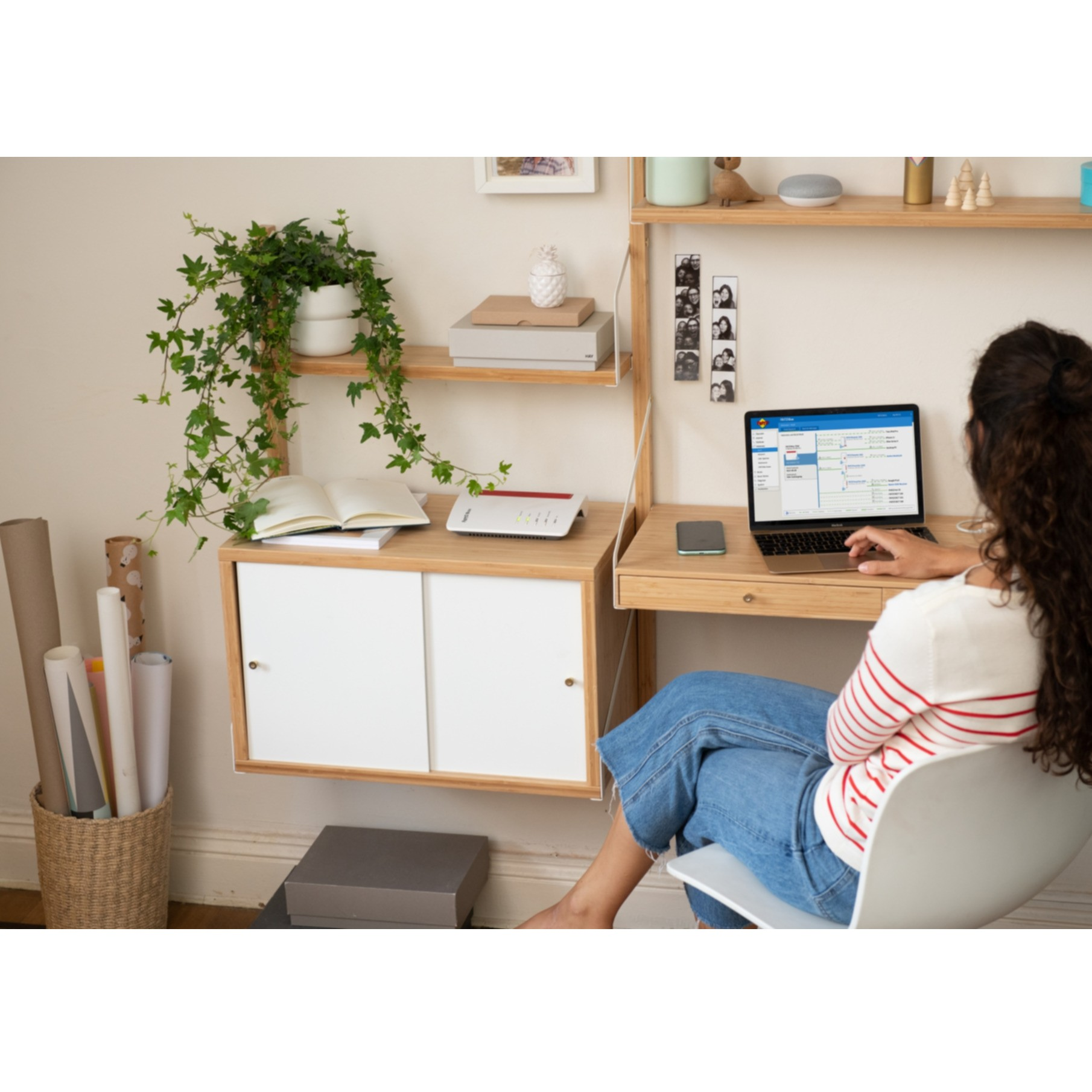 Kobieta pracuje na laptopie przy biurku z rośliną, książkami i routerem na półce. Jest okno.