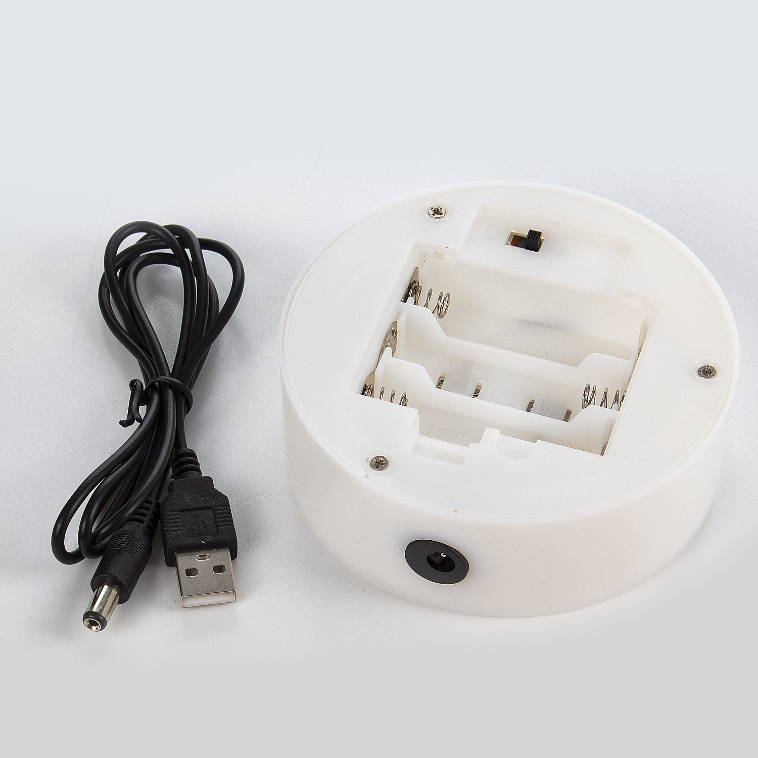 Base rotonda bianca con batterie, cavo USB e cavo di alimentazione nero. Sfondo bianco.