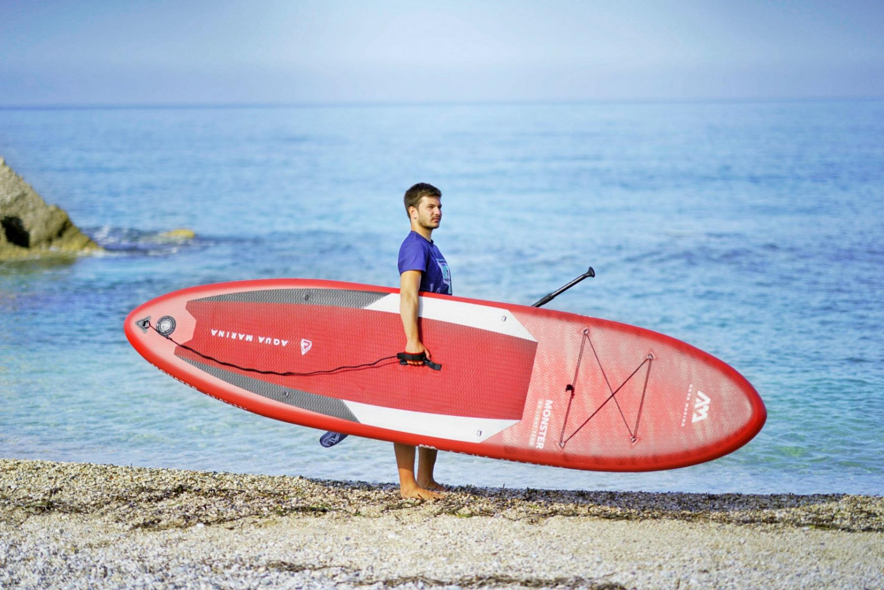 Ein Mann trägt ein rotes Paddleboard an einem Strand, das Meer im Hintergrund.