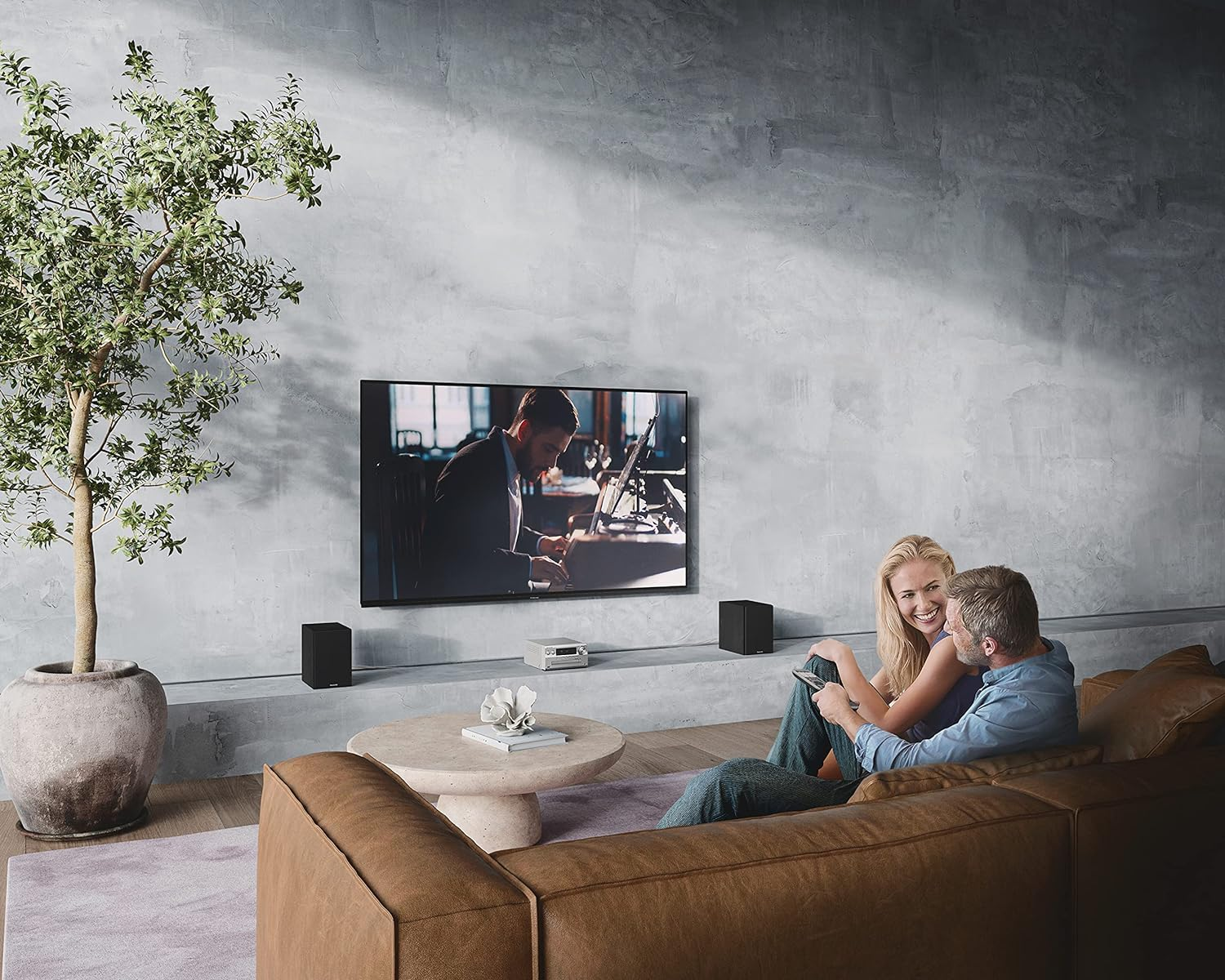 Couple assis sur un canapé marron, regardant la télé. Une plante et des enceintes dans la pièce.