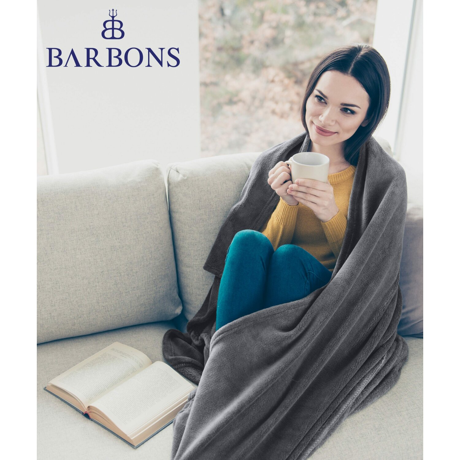 Frau in graue Decke gehüllt, hält Becher, liest Buch, sitzt auf Sofa.