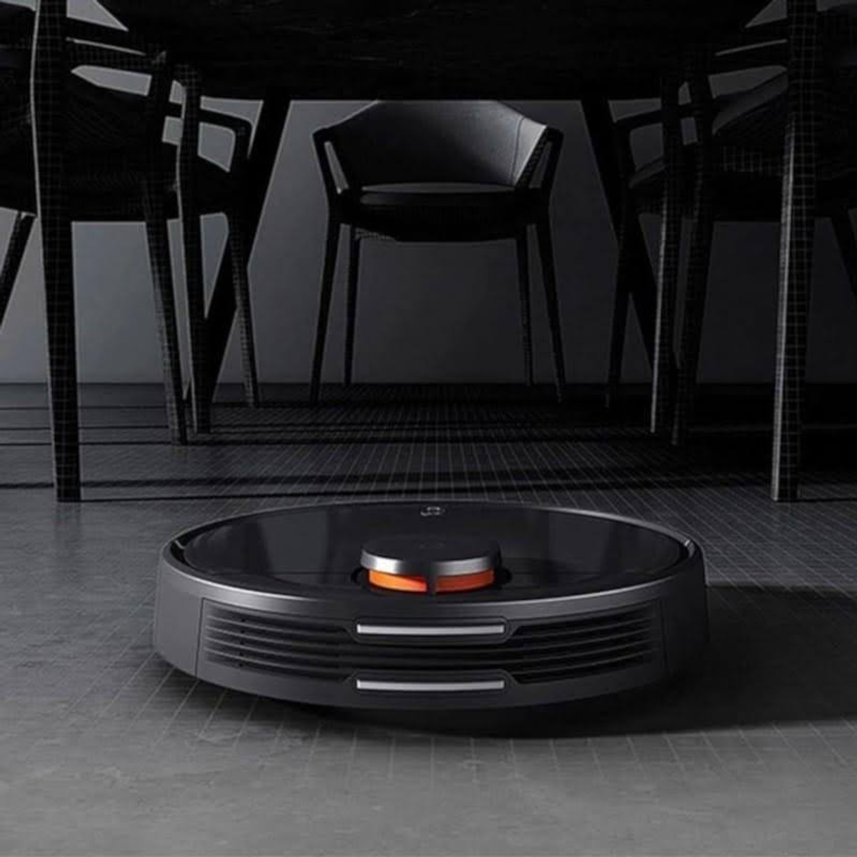 Aspiradora robot negra en un suelo gris debajo de una mesa y sillas. Ambiente oscuro.