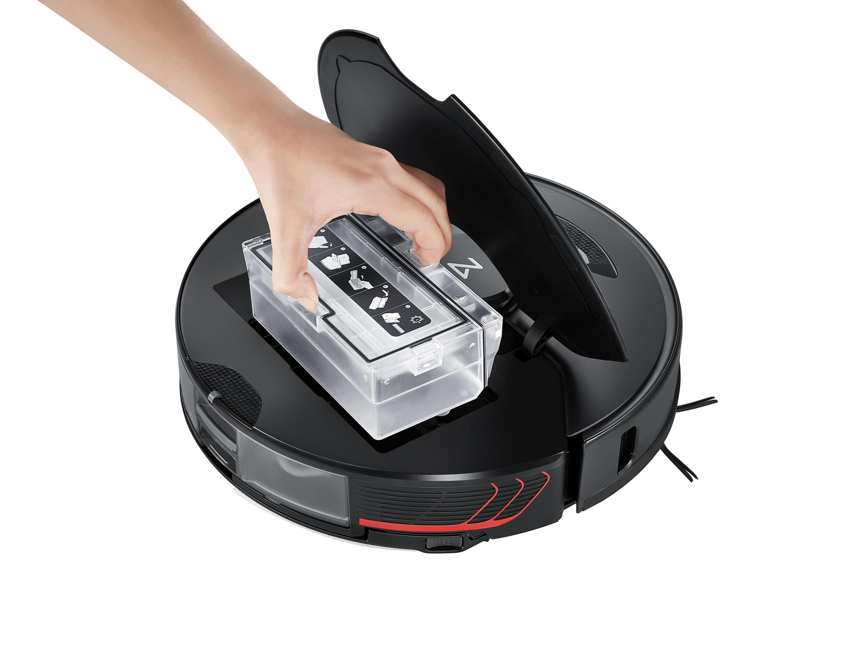 Robot odkurzający, czarny z czerwonym paskiem, dłoń wkładająca pojemnik, białe tło.