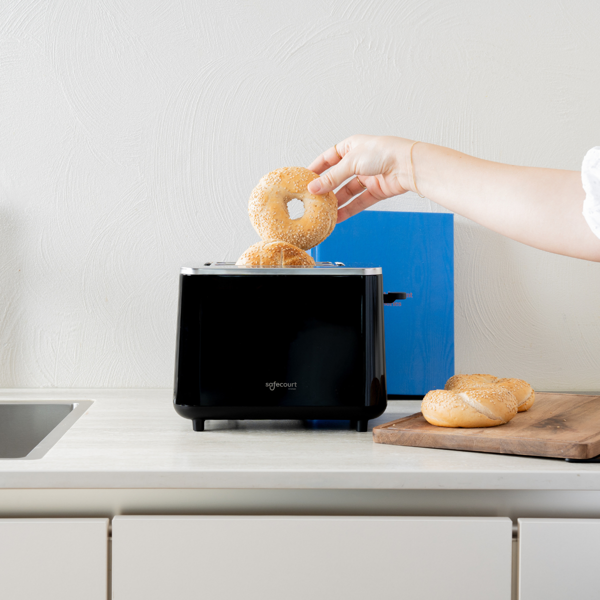 Hand die een bagel in een zwarte broodrooster legt. Meer bagels op een houten plank zijn zichtbaar.
