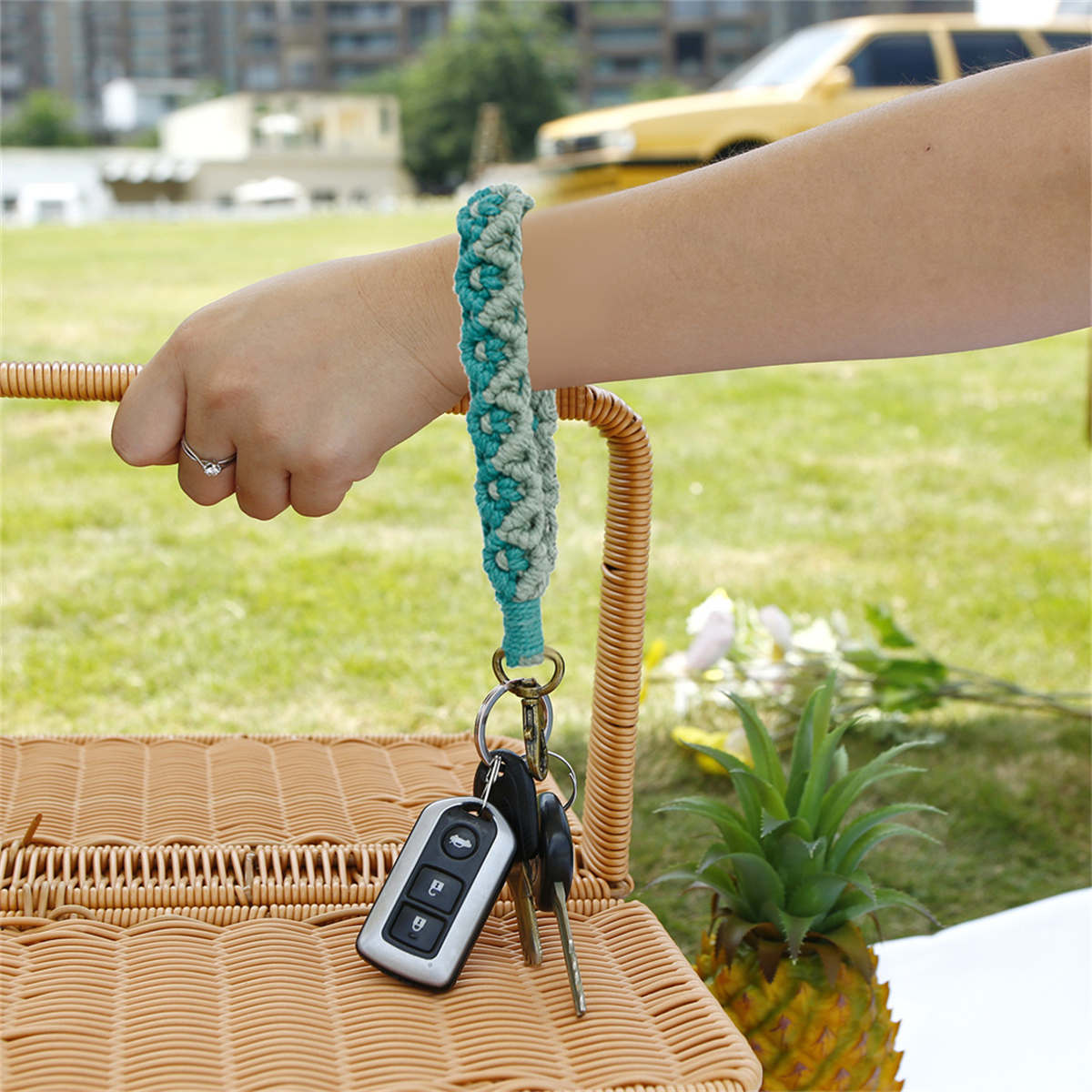 Eine Hand hält Schlüssel an einer türkisfarbenen und grauen Makramee-Handgelenkschlaufe. Sonnige Szene mit Gras und gelbem Auto.