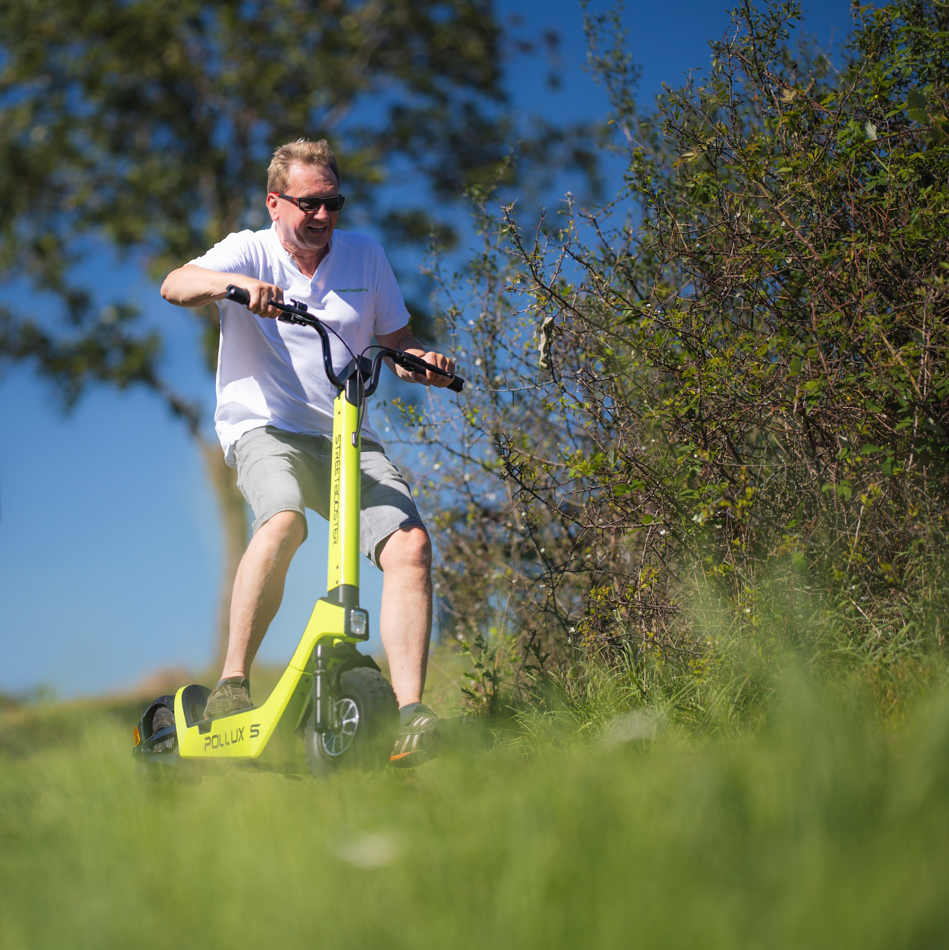 Mann auf gelbem Roller auf einem grasbewachsenen Hügel, Bäume und Himmel im Hintergrund.
