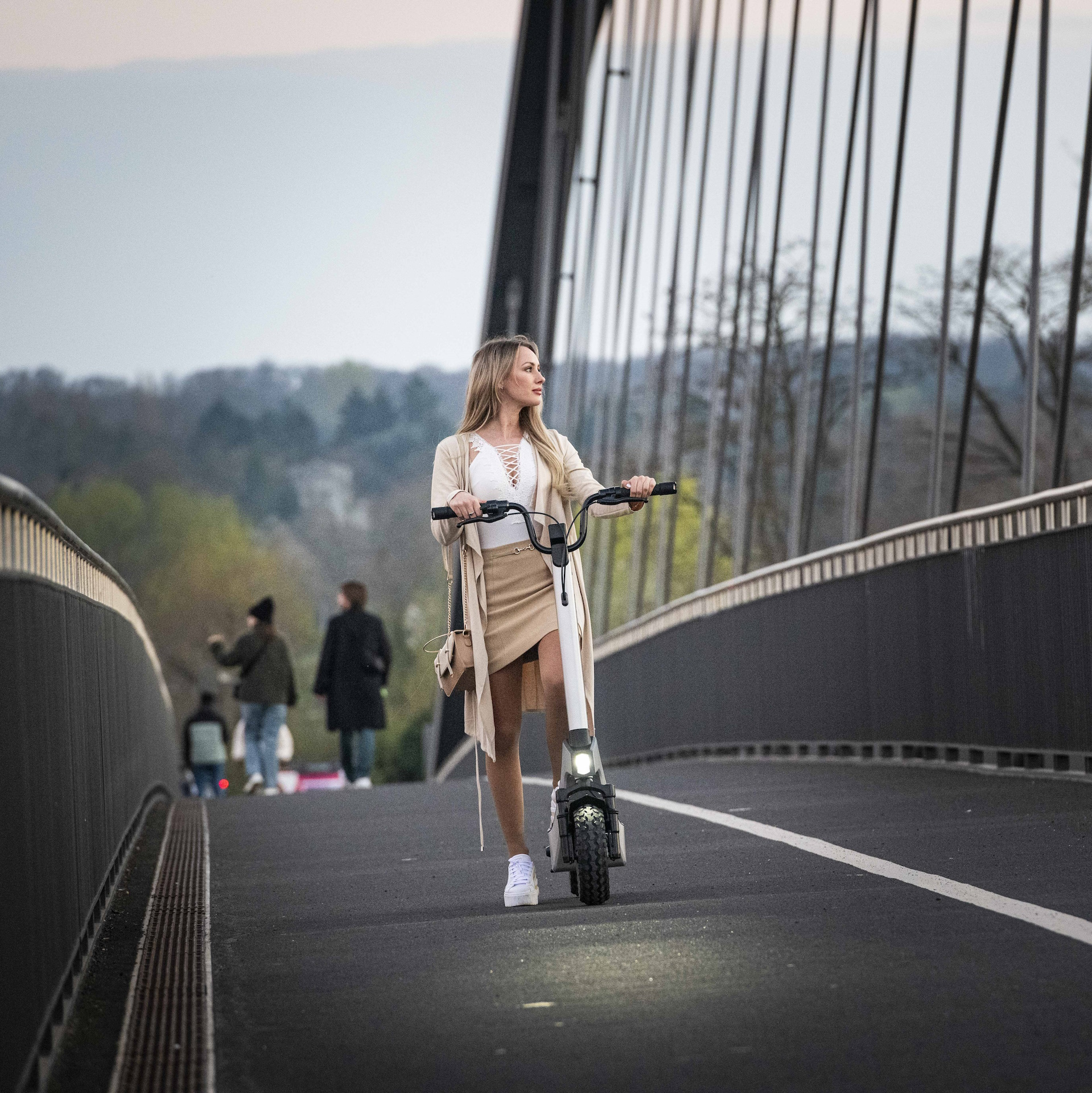 Frau fährt E-Scooter auf Brücke. Andere gehen im Hintergrund. Die Szene spielt sich im Freien ab.