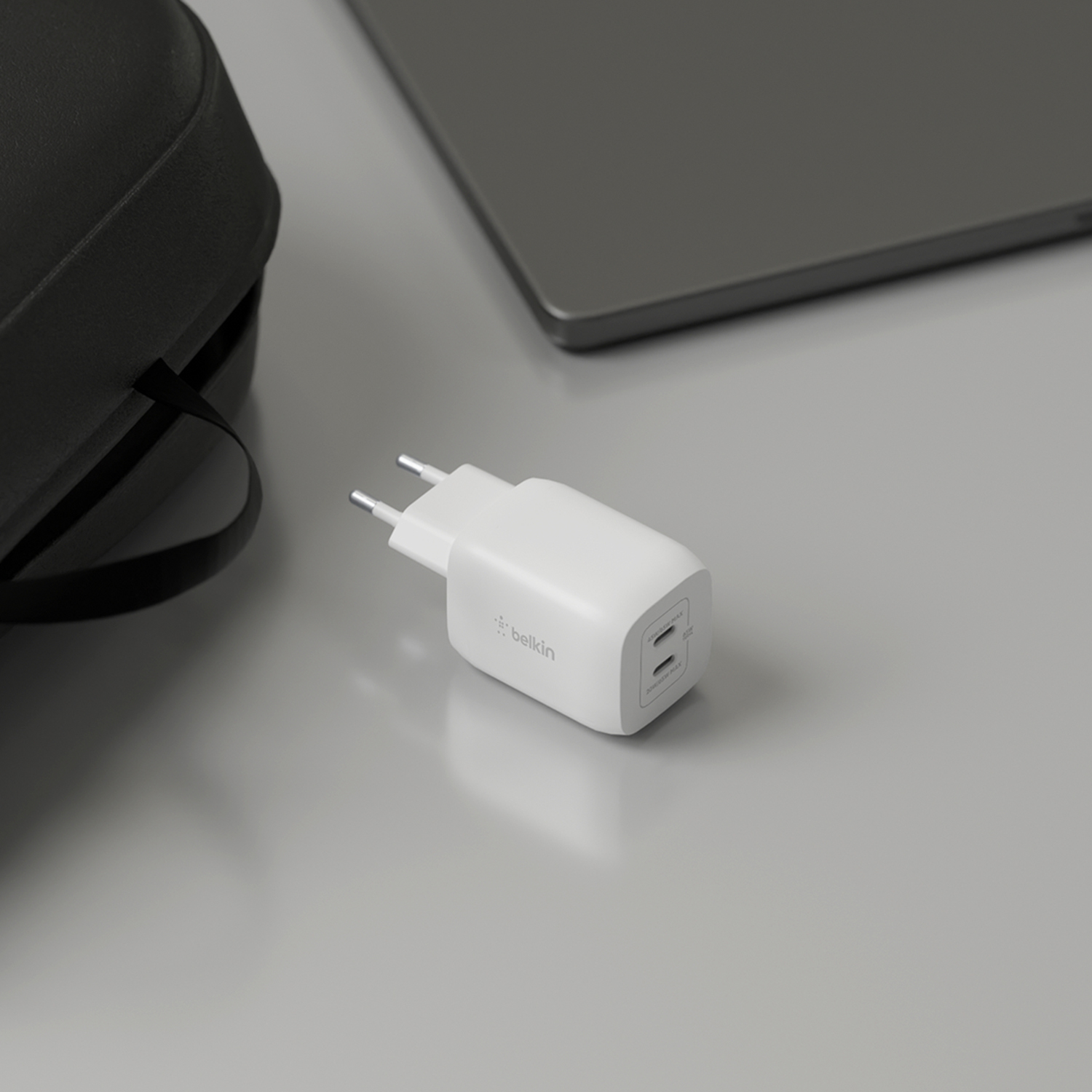 Caricatore USB-C Belkin bianco con due porte, accanto a una borsa nera e un laptop.