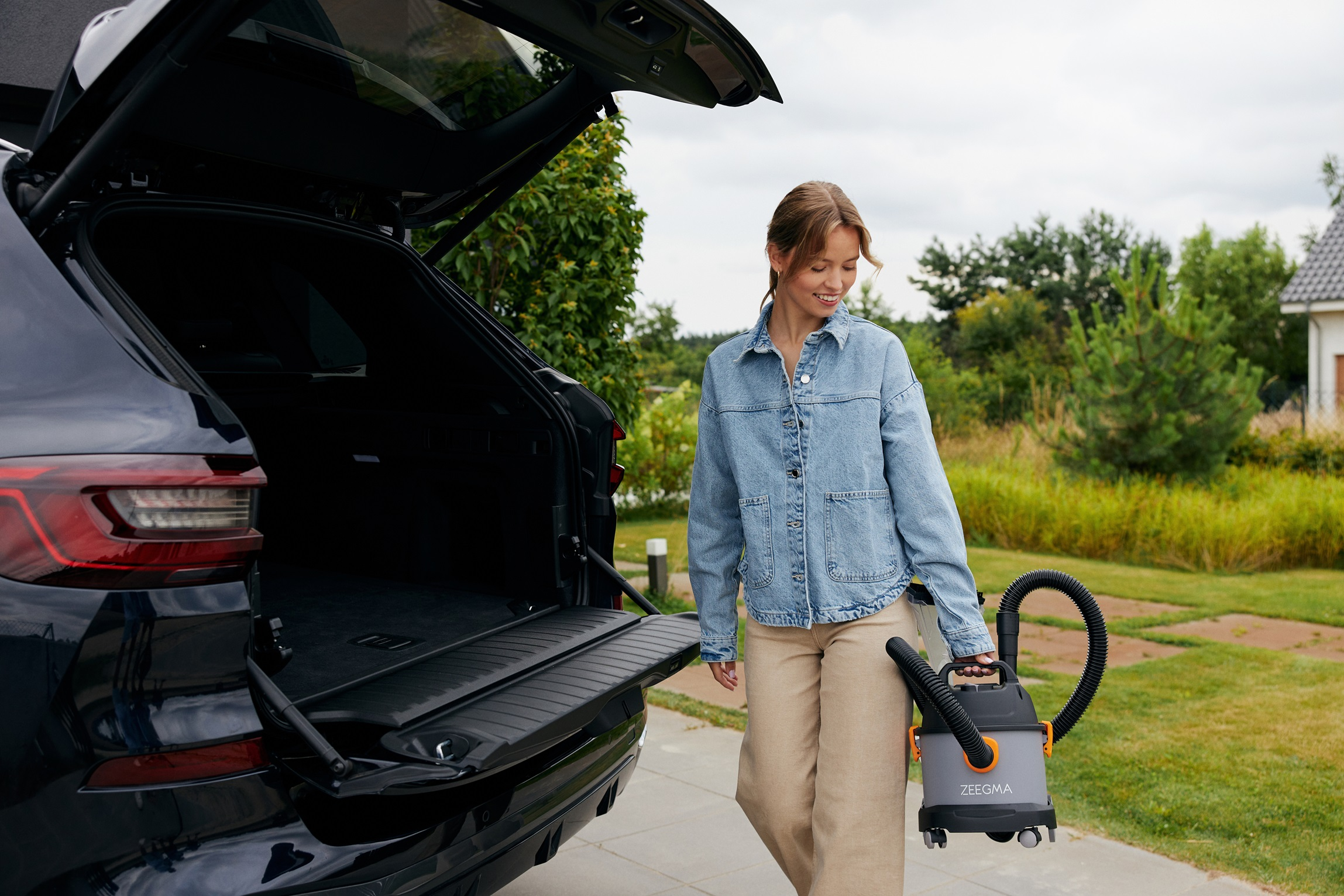 Kobieta trzyma odkurzacz w pobliżu otwartego bagażnika samochodu. Zielona trawa i drzewa w tle.