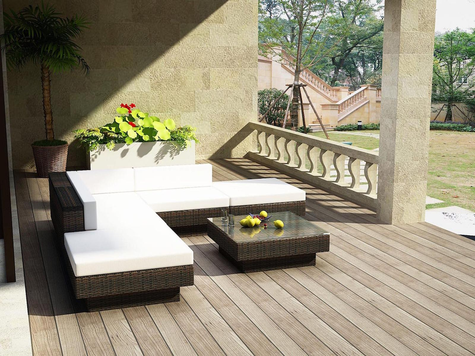 Terrassenset mit weißen Kissen, braunem Korbgeflecht und einem Tisch mit Glasplatte. Palme und Grün im Hintergrund.