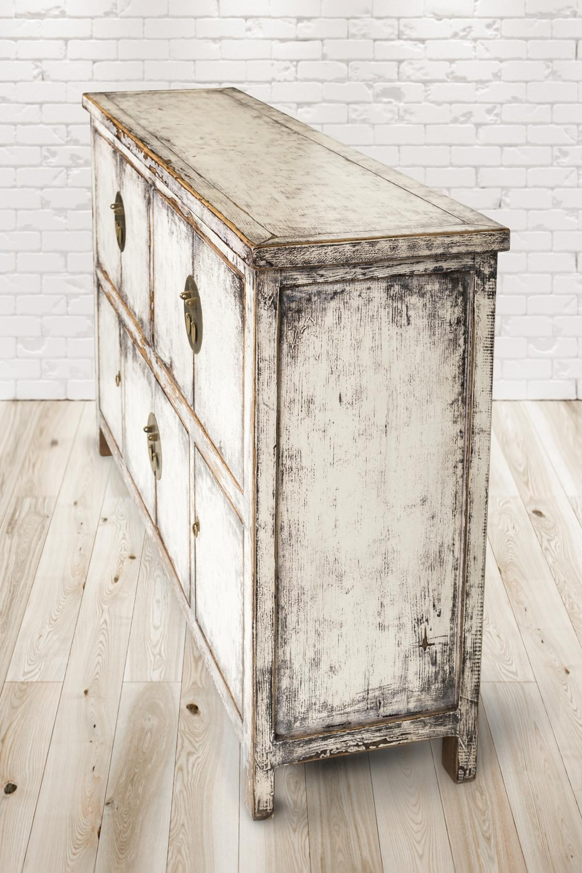 Ein weißer, abgenutzter Holzschrank mit vier Schubladen, Messinggriffen, auf hellem Holzboden.