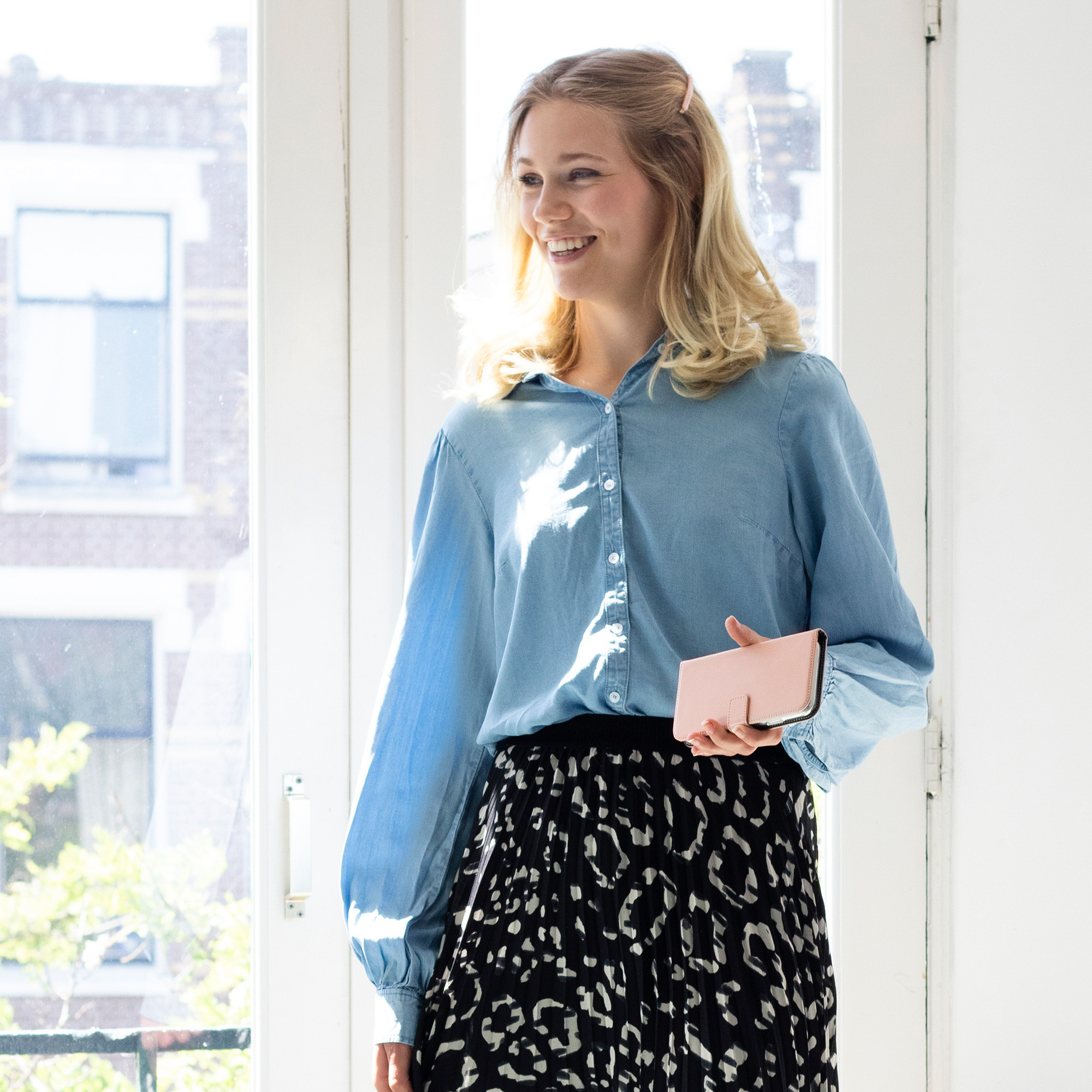 Femme blonde souriante tenant un portefeuille rose. Elle porte une chemise bleue et une jupe noire à motifs. Fenêtre claire.