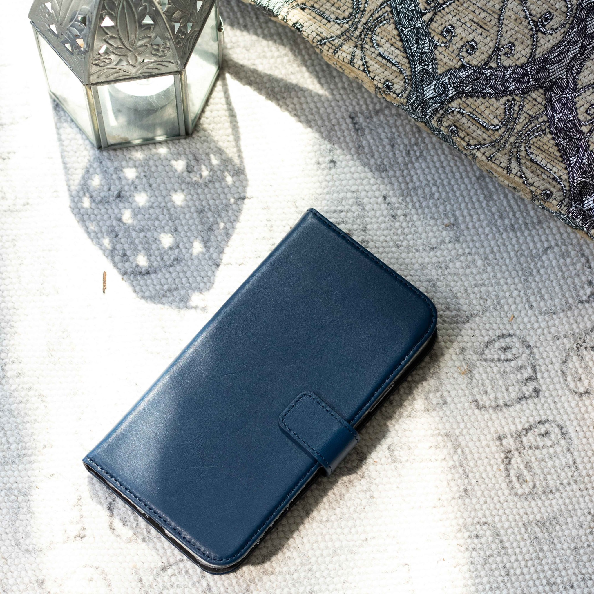Étui de téléphone bleu avec fermeture à bouton sur un tapis blanc. Lampe argentée et oreiller en arrière-plan.