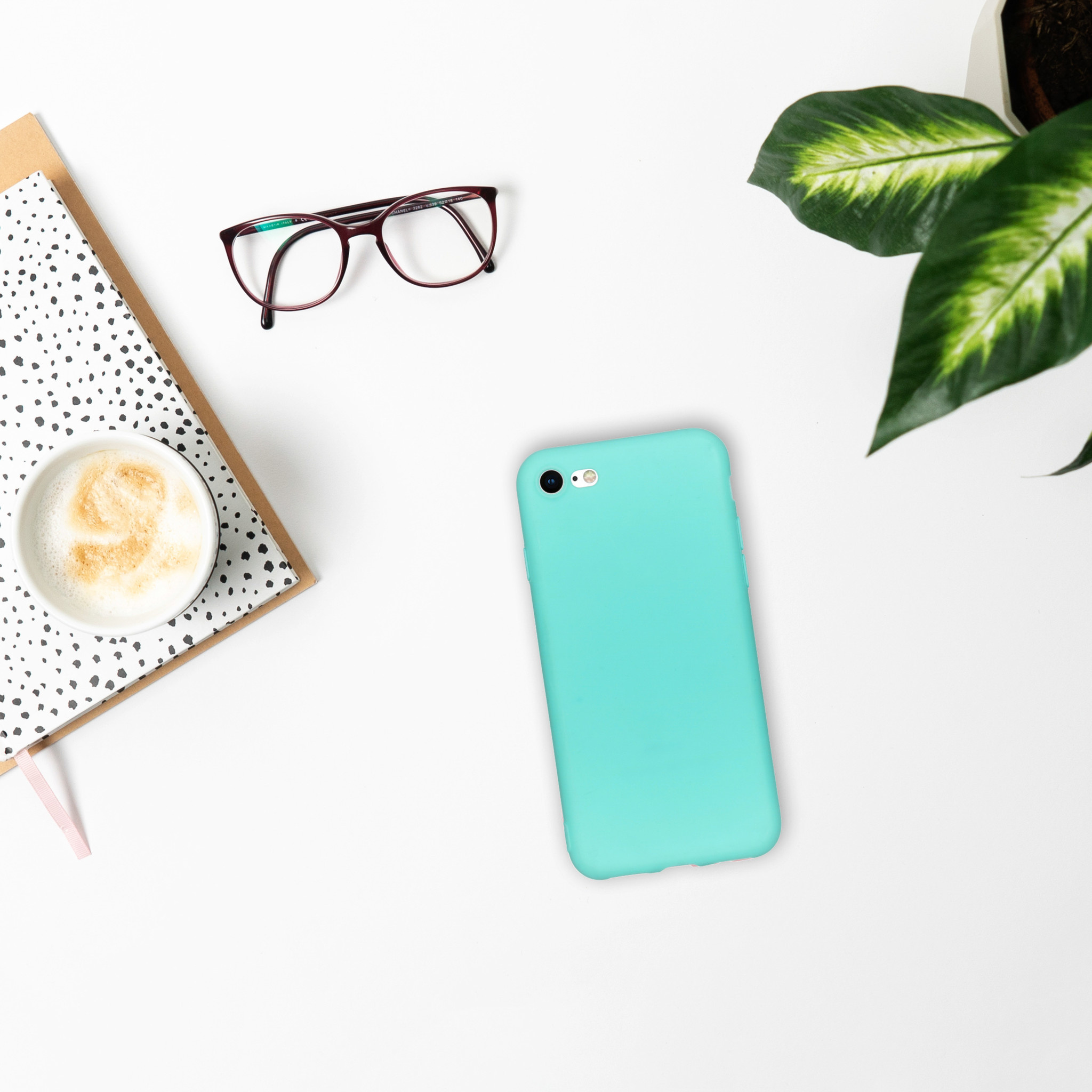 Téléphone dans un étui sarcelle, sur une surface blanche. À côté, des lunettes, un bloc-notes et une tasse de café.