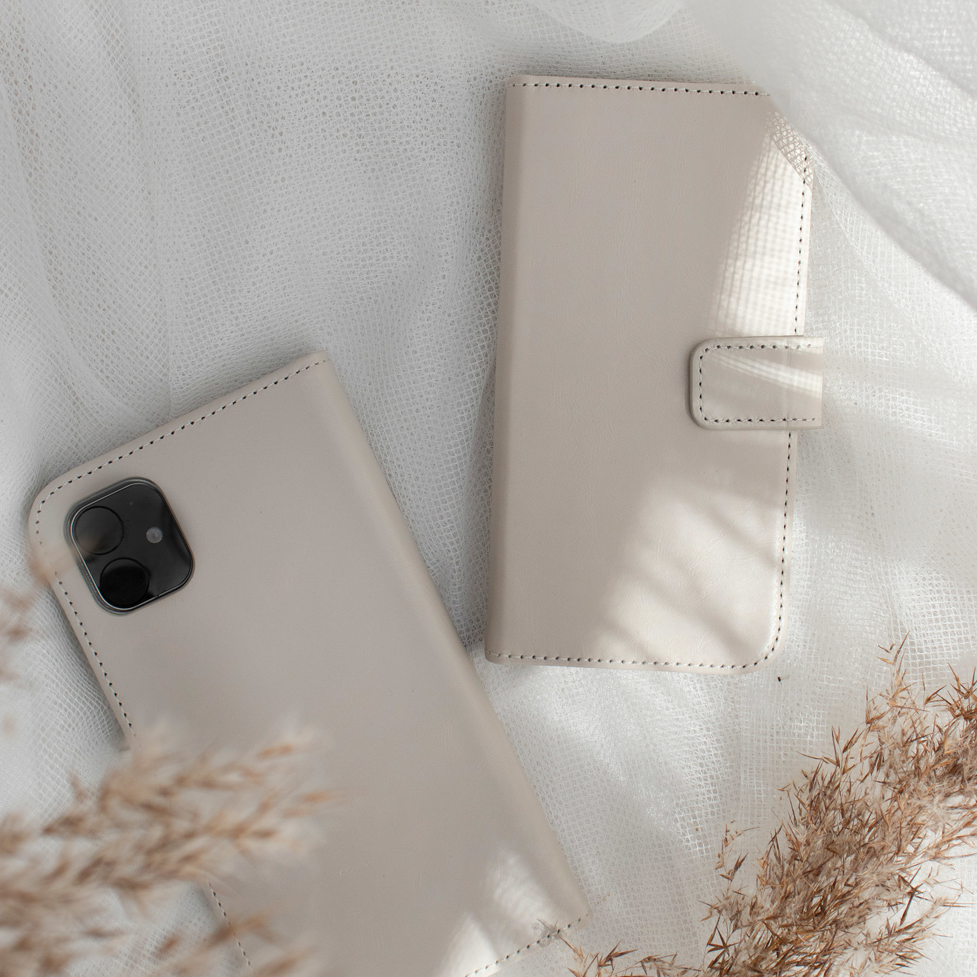 Deux étuis de téléphone beiges sur un tissu blanc. Un ouvert, un fermé. Plantes séchées en arrière-plan.