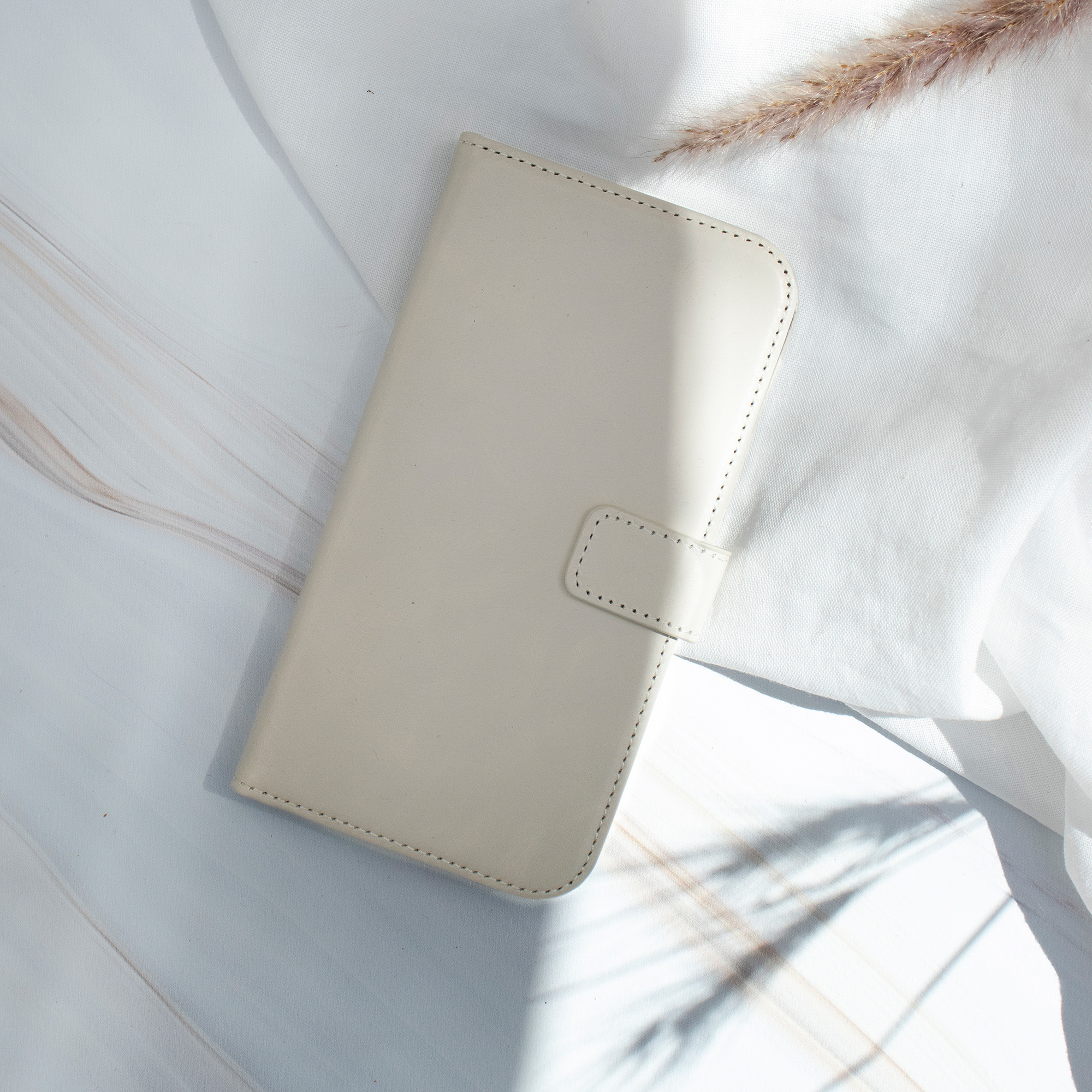 Coque de téléphone beige, placée sur un tissu blanc avec une plante sèche.