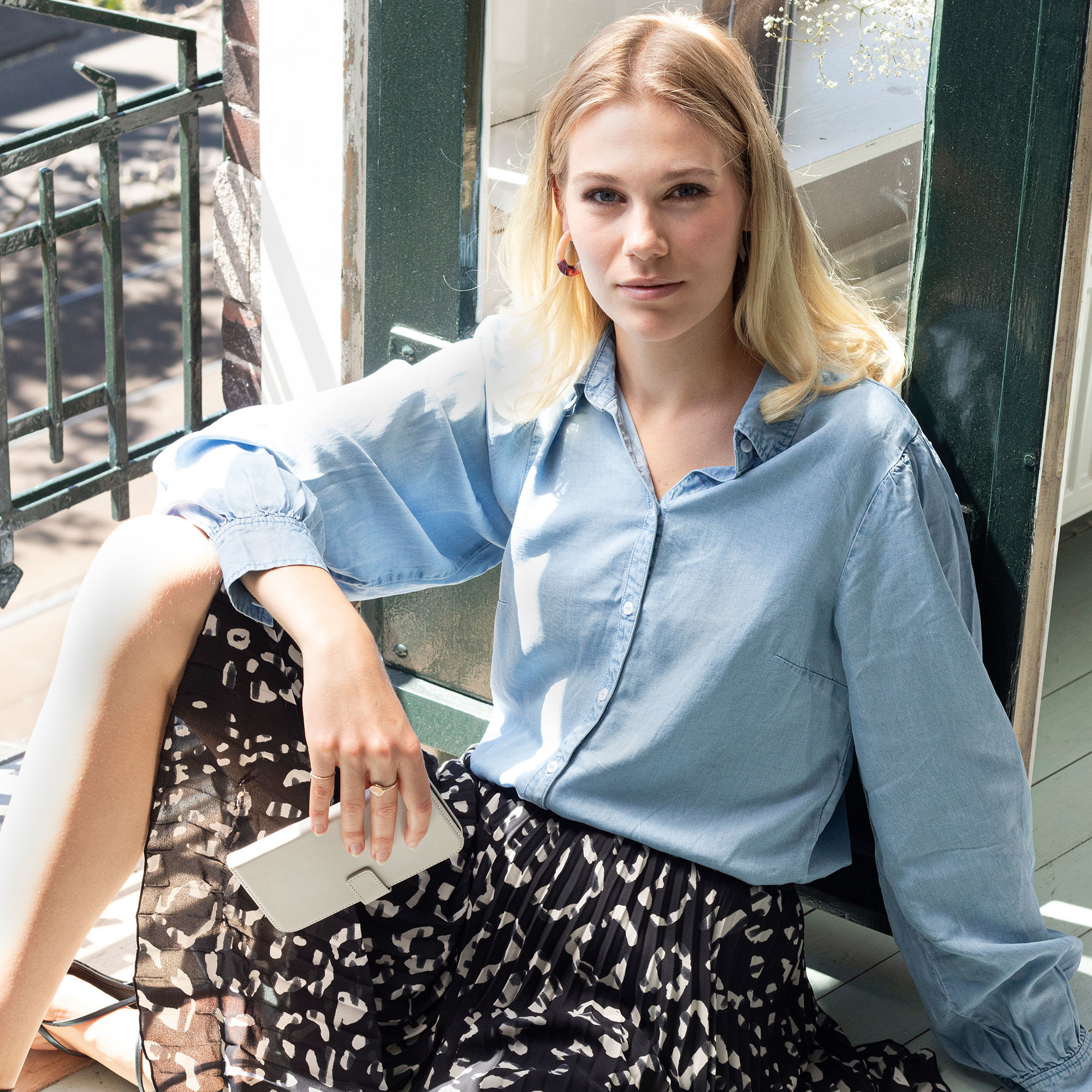 Femme sur un balcon, portant une chemise bleue et une jupe, tenant une coque blanche.