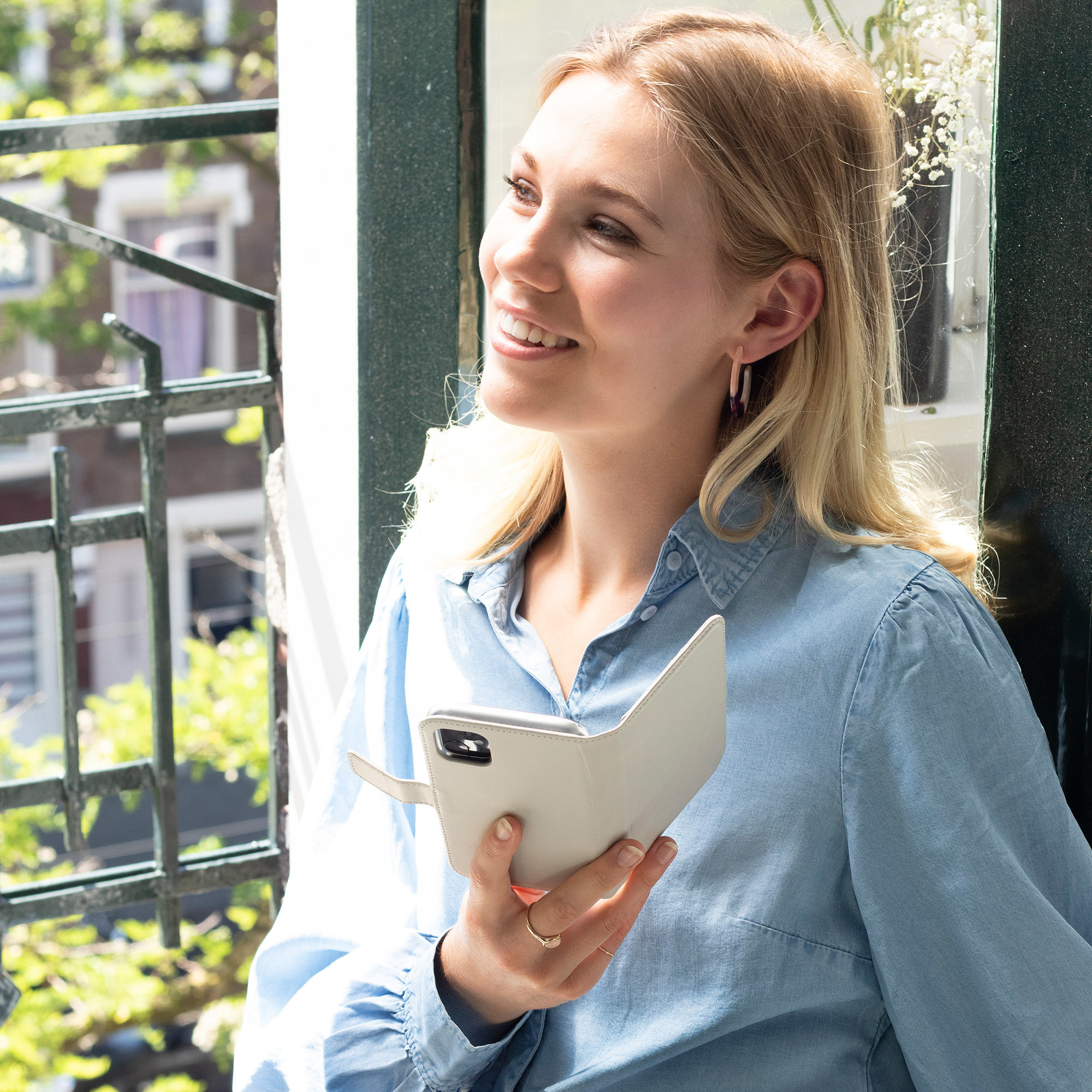 Femme regardant par une fenêtre, tenant un étui de téléphone. Elle sourit. L'étui est blanc.