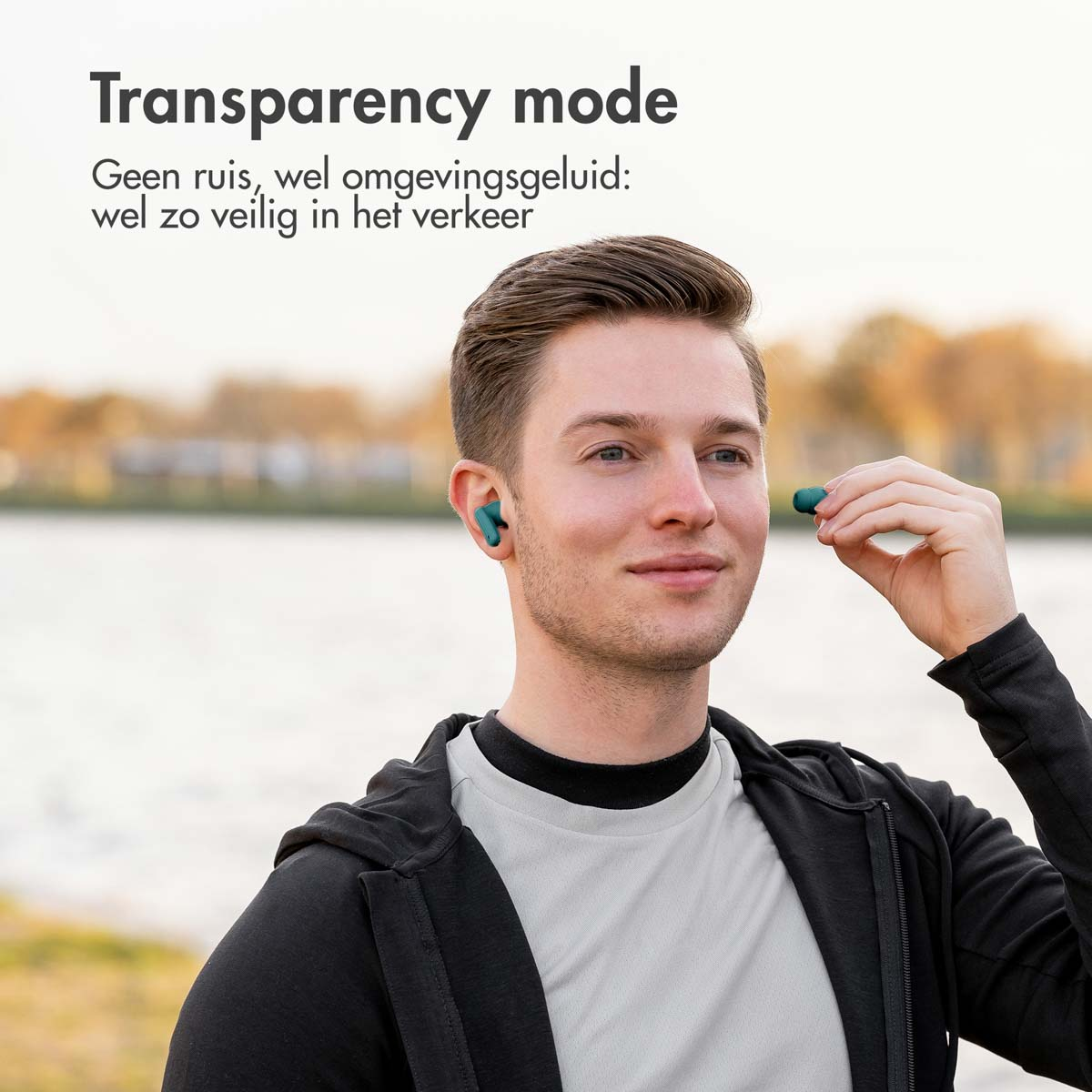 A man wearing earbuds. The man is smiling, and is in a park setting.