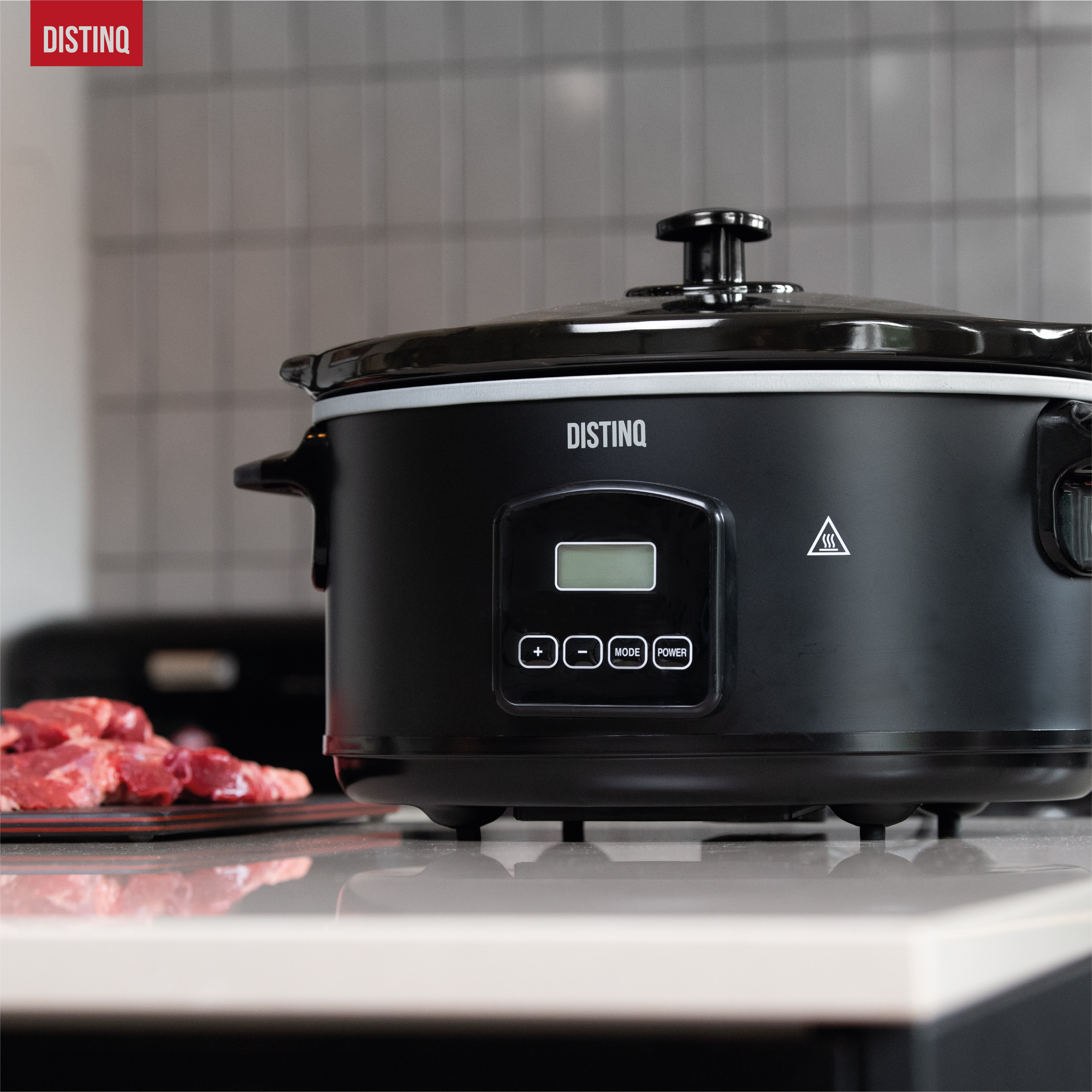 Zwarte slowcooker met bediening, deksel en handvat op een aanrecht. Vlees ligt op een snijplank.