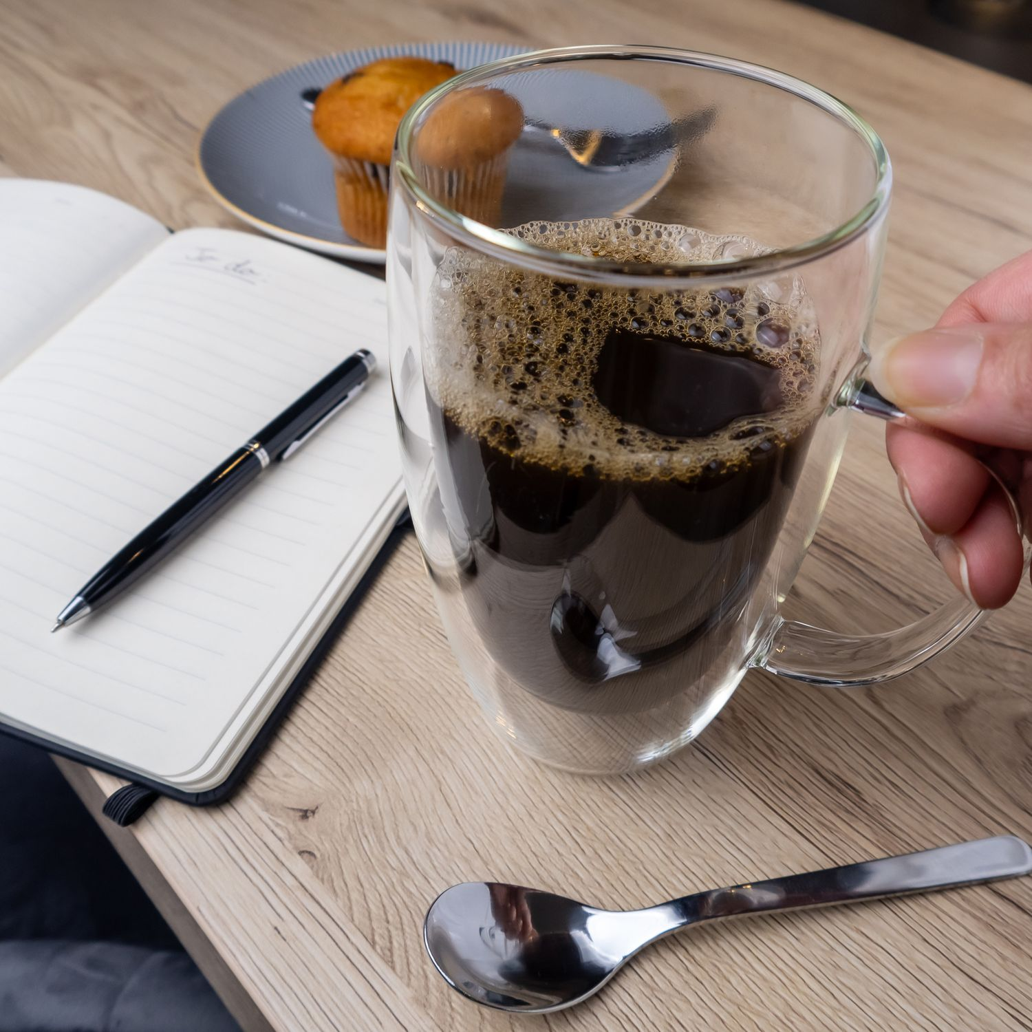 Nahaufnahme einer Glastasse mit Kaffee, Stift, Notizbuch und Muffin auf einem Teller.