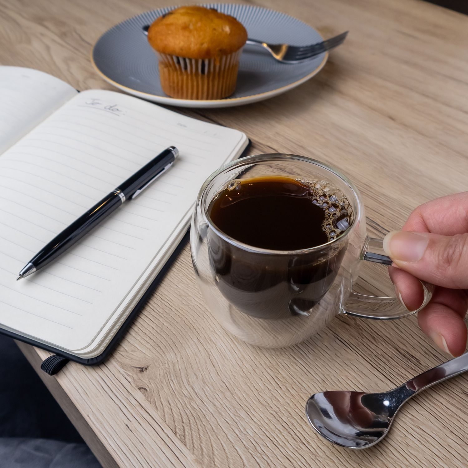 Eine Kaffeetasse, ein Notizbuch, ein Stift und ein Muffin auf einem Holztisch.