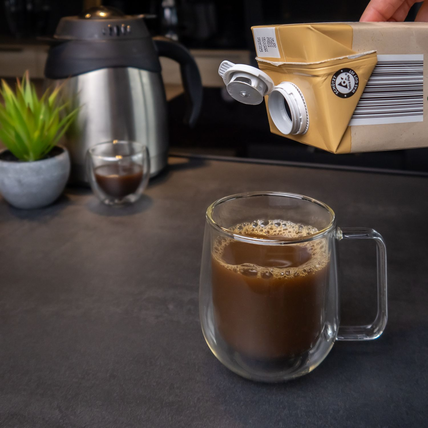 Kaffee in einem Glasbecher, der aus einem braunen Karton gegossen wird. Eine Pflanze ist im Hintergrund.