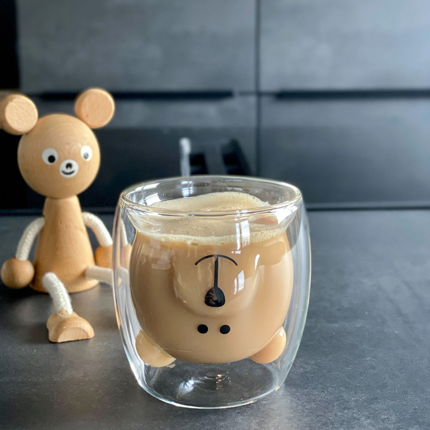 Bärenförmige Kaffeetasse mit Schaum. Holz-Bärenspielzeug daneben auf dunkler Oberfläche.