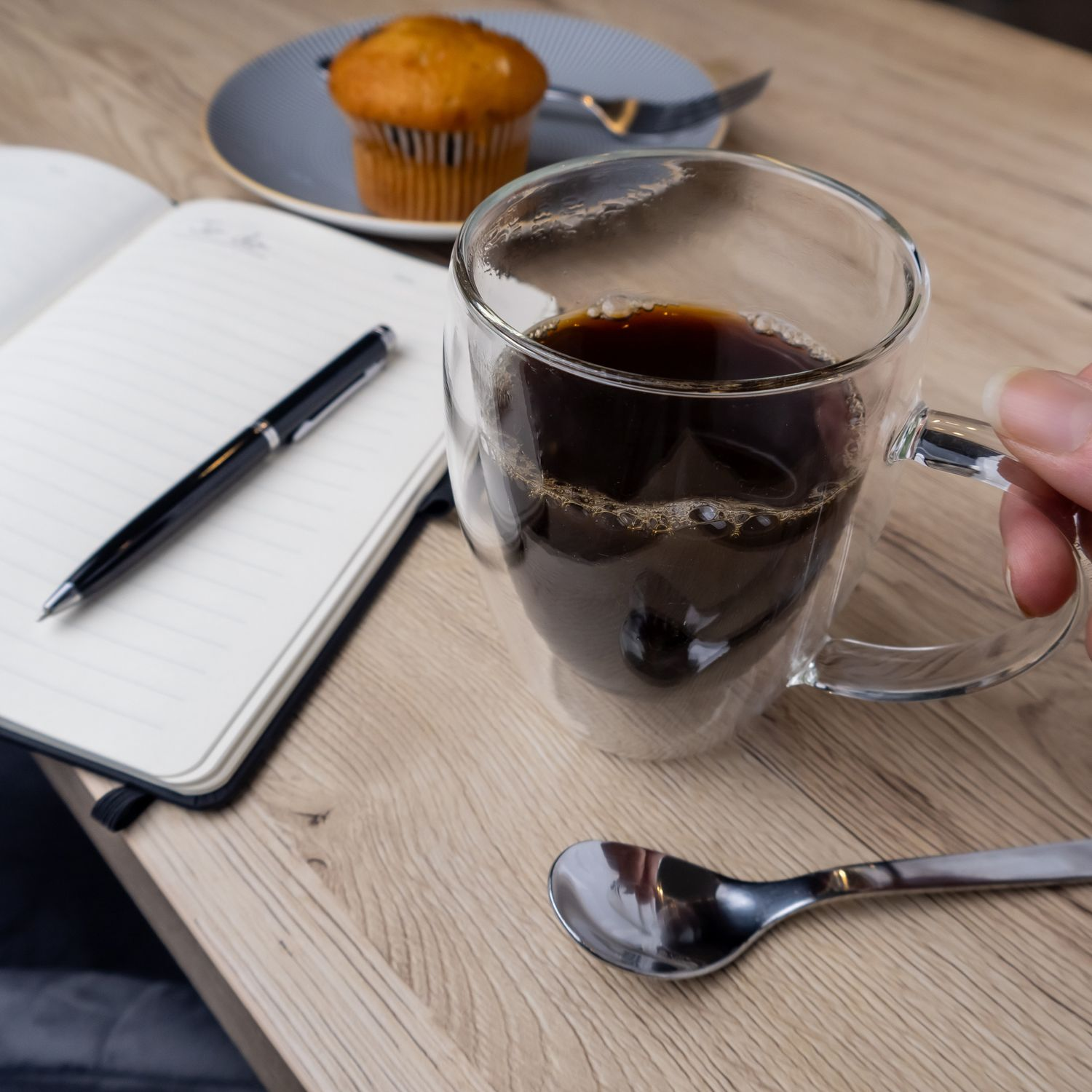 Kaffeetasse, Notizbuch, Stift und ein Muffin auf einem Holztisch. Hand hält Tasse.