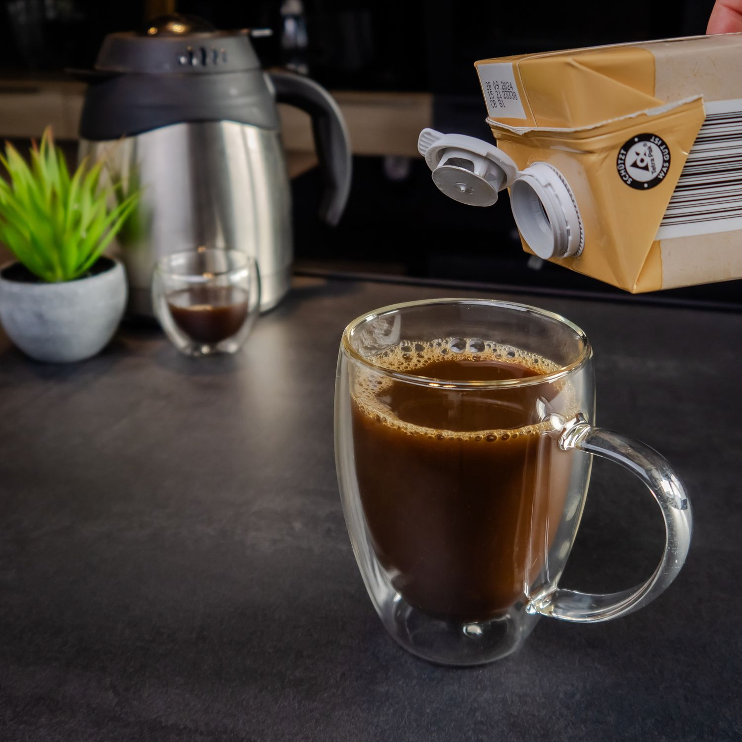 Eine Tasse Kaffee in einem Glasbecher, ein Karton und eine Pflanze auf dunkler Theke.