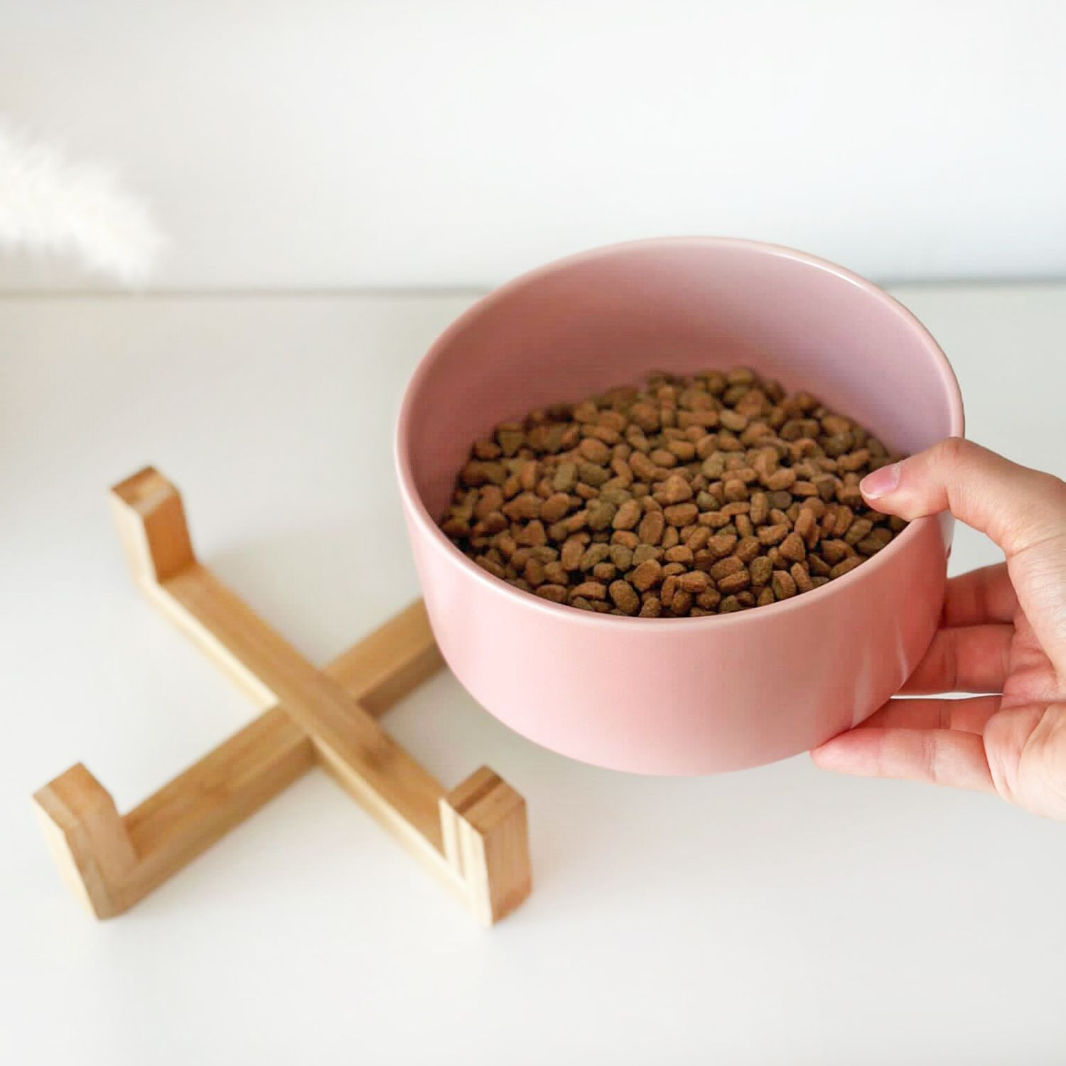 Eine rosa Schüssel mit Trockenfutter auf einem Holzständer, gehalten von Hand.