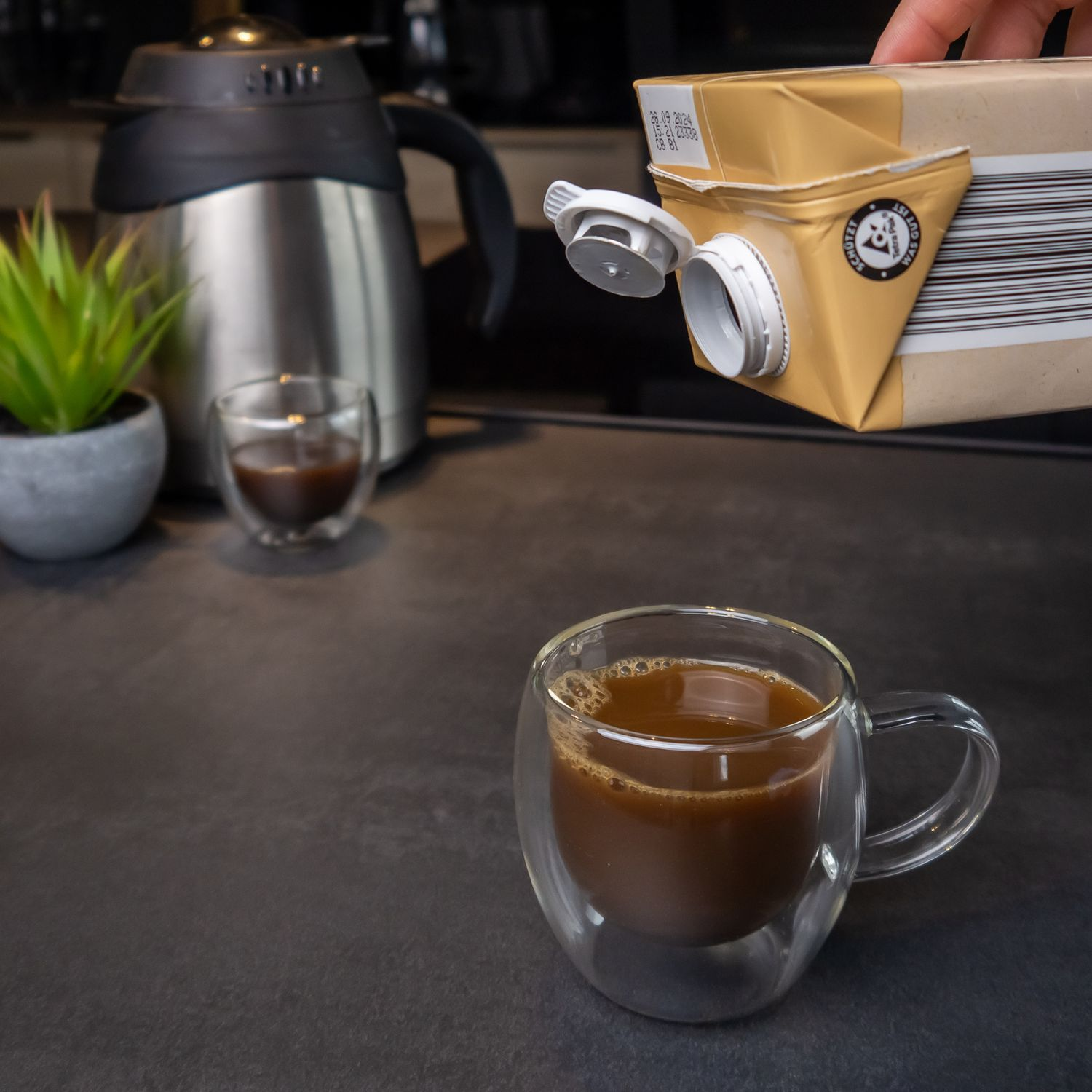 Kaffee aus einem Karton in eine Glastasse gießen, mit kleiner Tasse im Hintergrund.