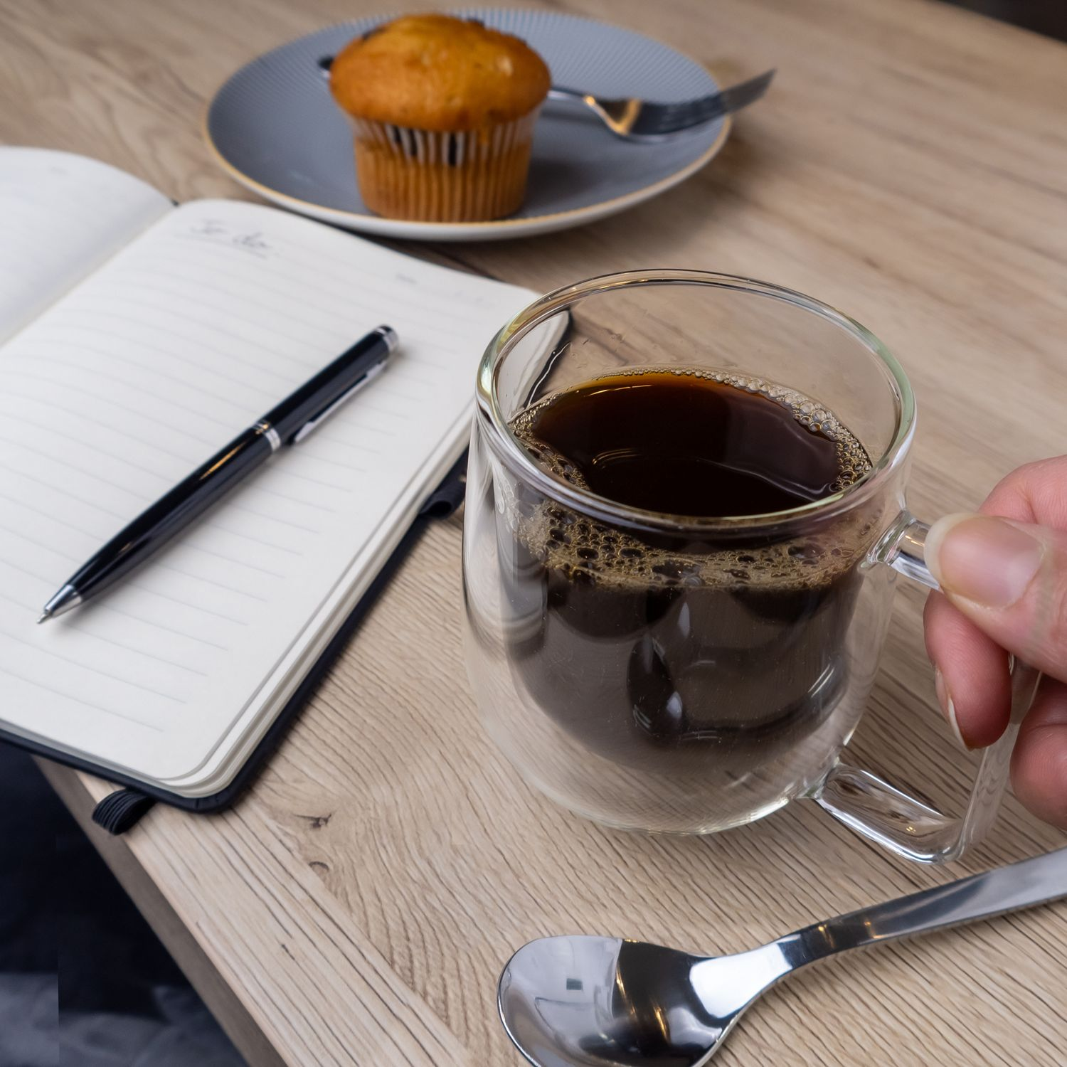 Eine Kaffeetasse, ein Stift, ein Notizbuch, ein Muffin und ein Löffel auf einem Holztisch. Hand hält die Tasse.