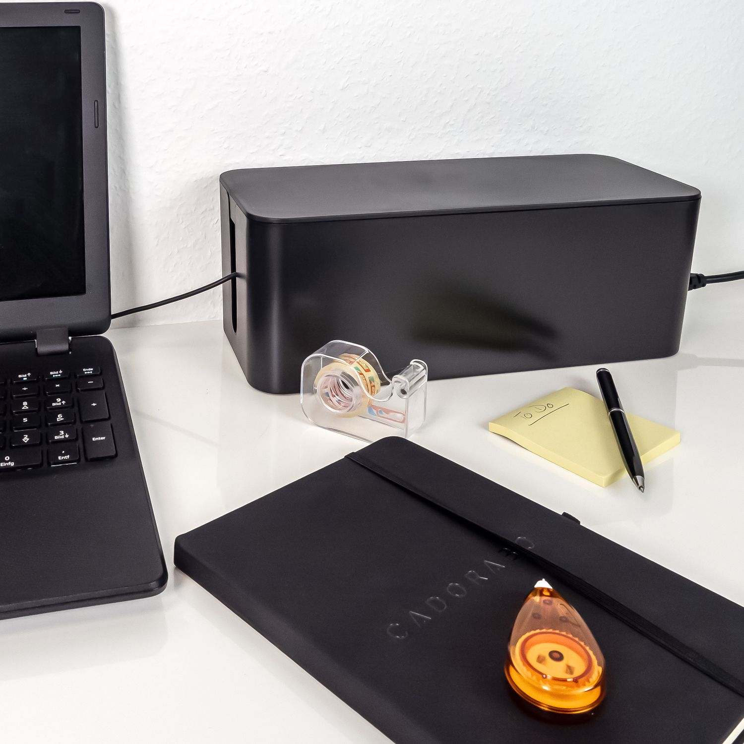 Schreibtisch-Setup mit Laptop, Klebebandabroller, Notizbuch und Stift.