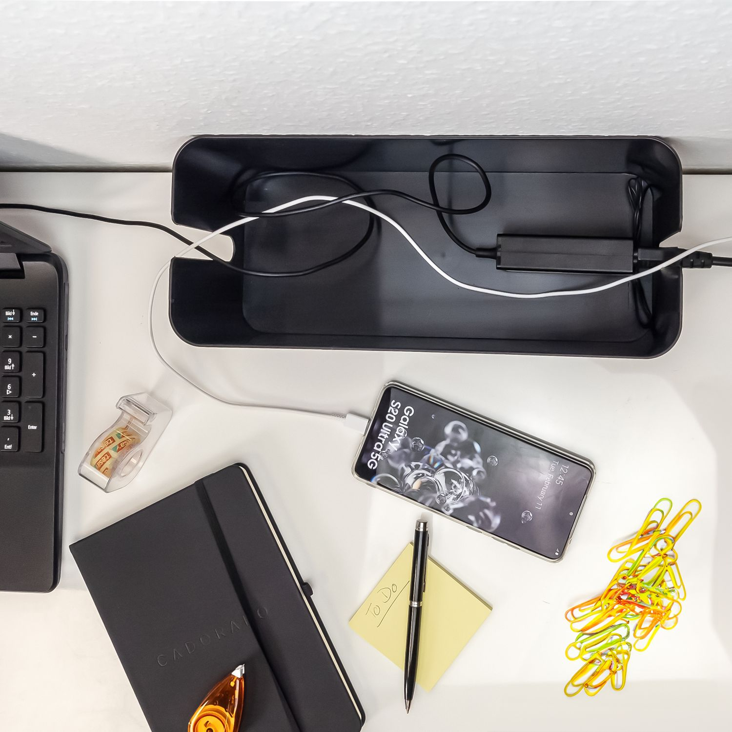 Schreibtischansicht mit Telefon, Notizbuch, Stift, Büroklammern und Kabeln in einem schwarzen Organizer.