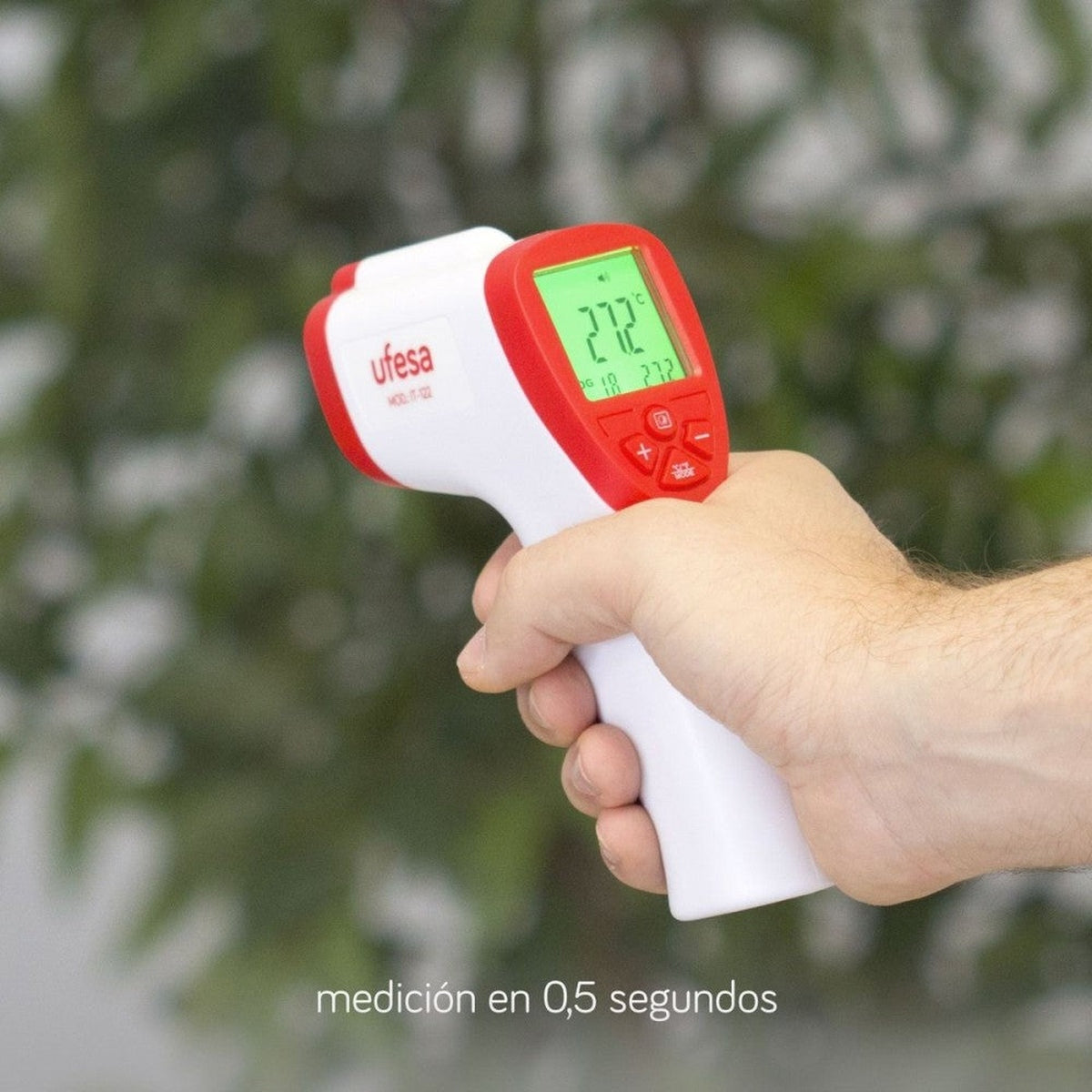 Mano sosteniendo un termómetro infrarrojo rojo y blanco, mostrando 27.2 grados. Fondo verde.