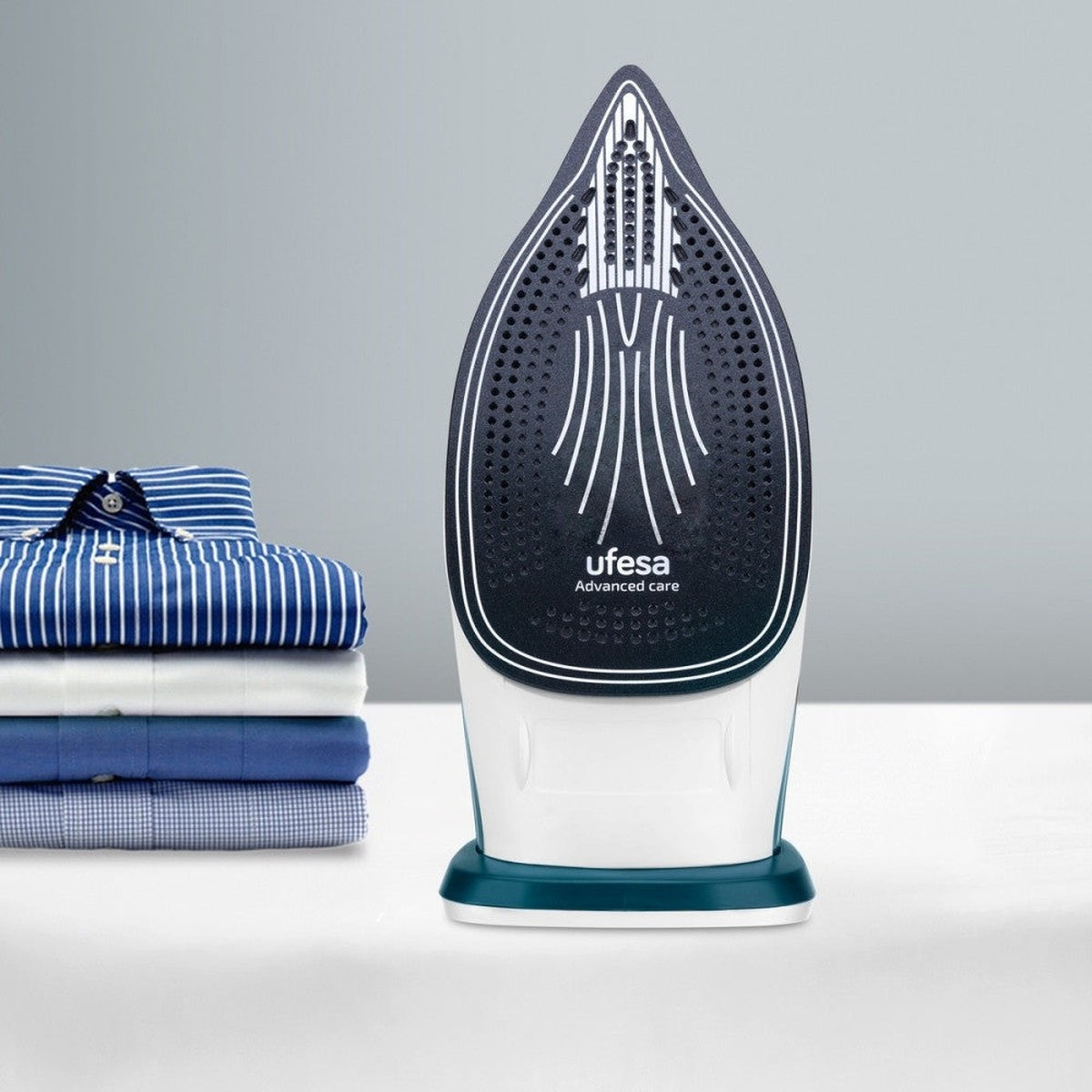 Una plancha con base azul y una pila de camisas dobladas. La plancha está en primer plano.