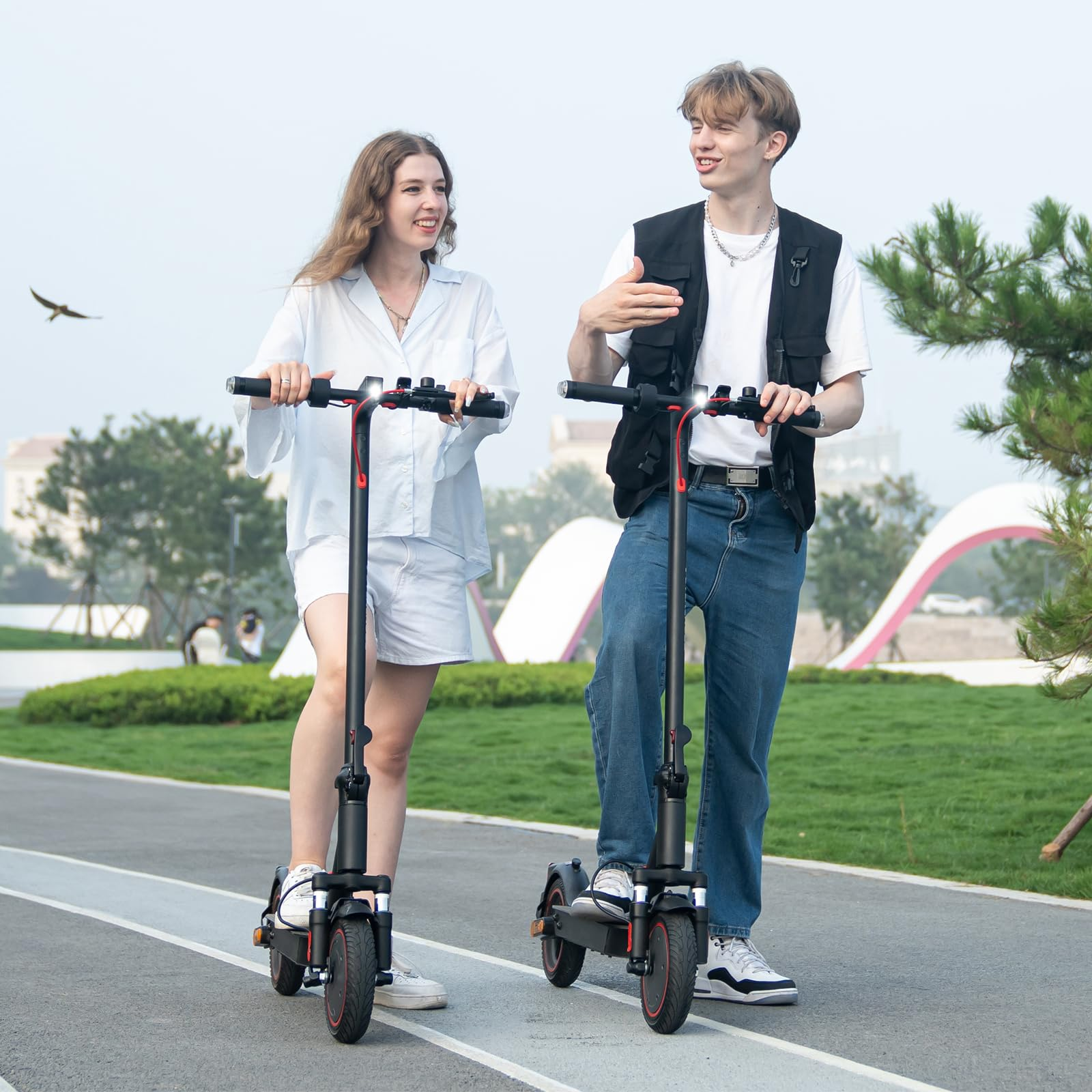 Twee mensen op scooters, lachend. Ze bevinden zich op een pad. De achtergrond is een park met bomen en gebouwen.
