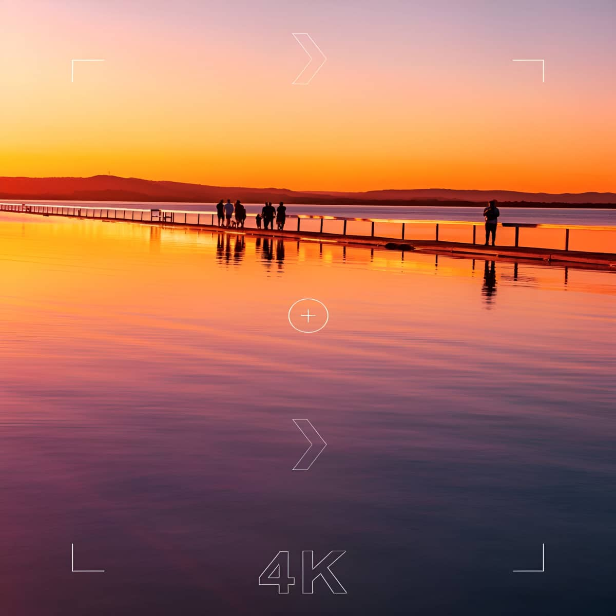 Sonnenuntergang über ruhigem See. Menschen auf einem Steg. Bunter Himmel spiegelt sich im Wasser. Berge im Hintergrund.