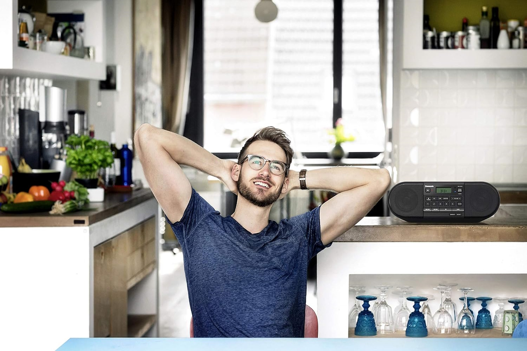 Uomo in cucina, sorridente con le mani dietro la testa. Sfondo cucina, bicchieri negli armadietti.