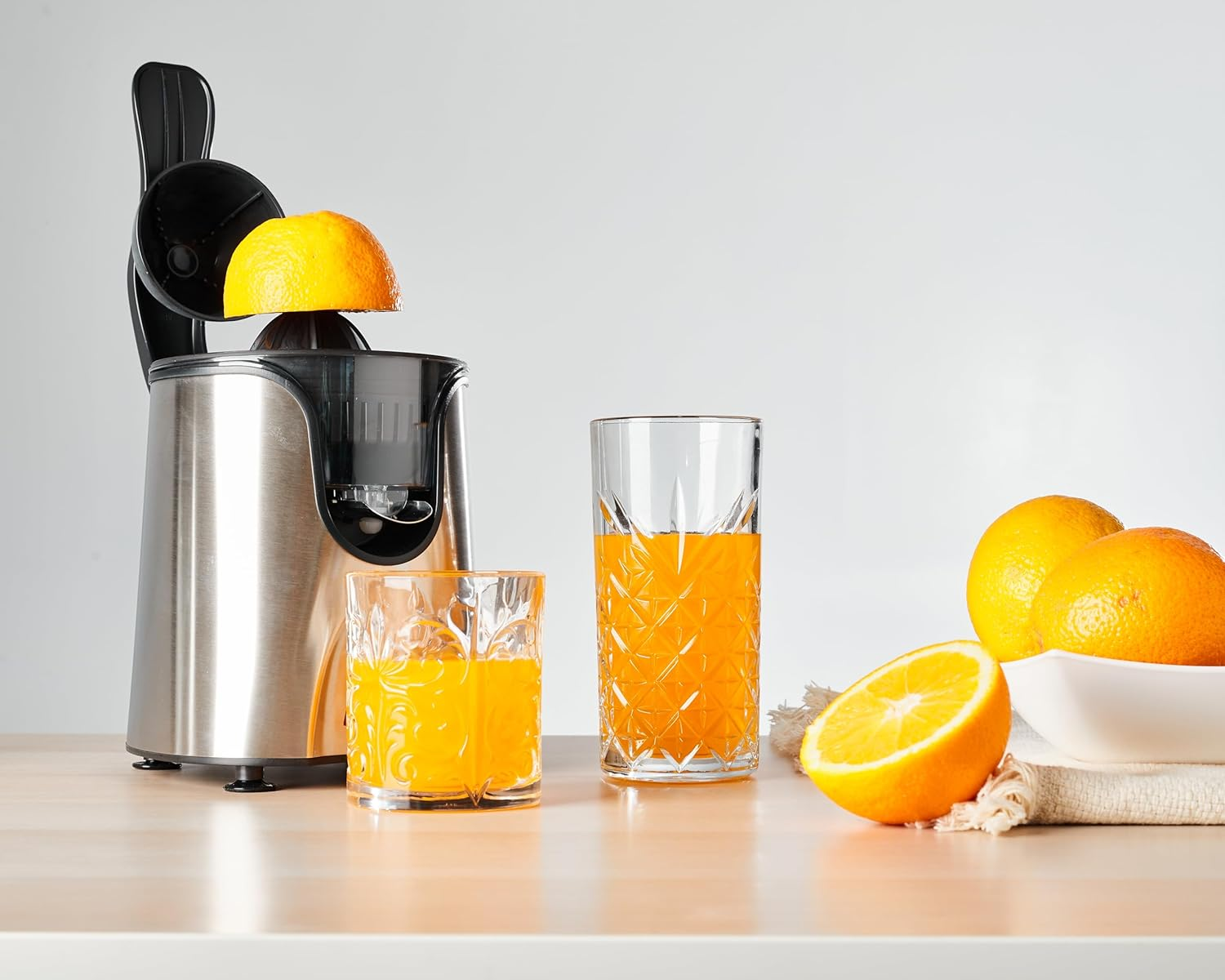Sokowirówka z przeciętą pomarańczą w trakcie wyciskania. Dwie szklanki soku i całe pomarańcze.