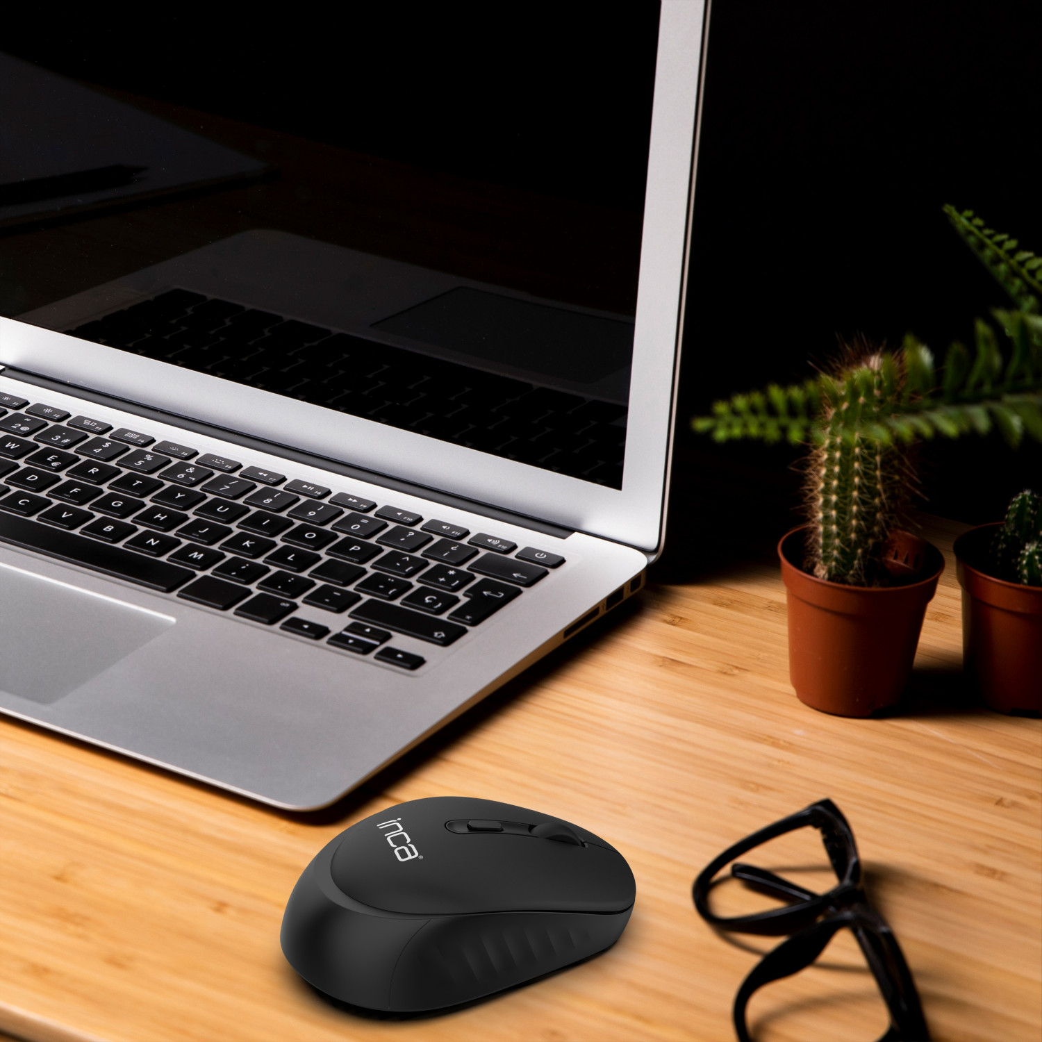 Schwarze kabellose Maus auf Holzschreibtisch, Laptop, Brille und Pflanzen sichtbar.