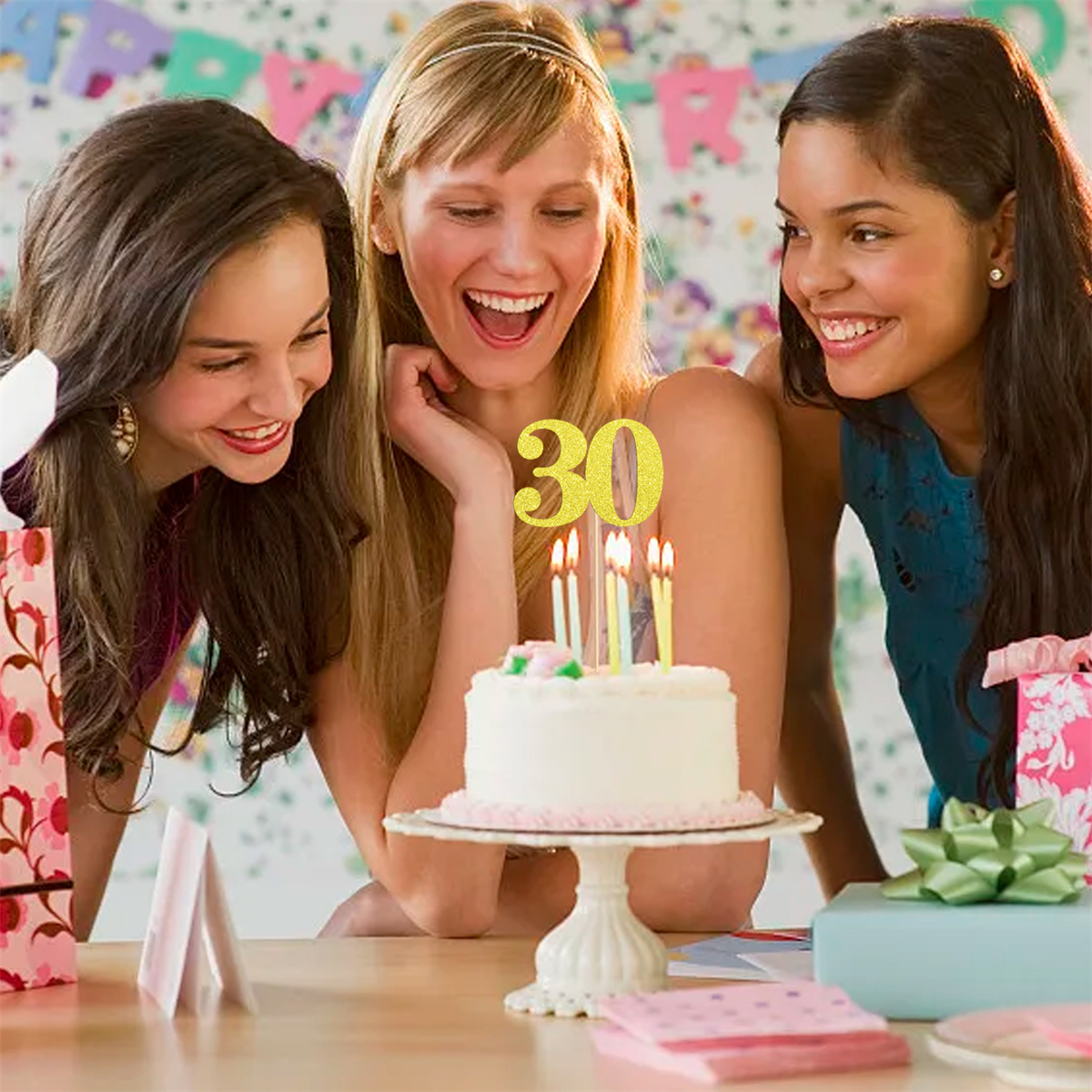 Drei Frauen lächeln um eine Torte mit einer goldenen "30" Kerze auf einem Tisch.