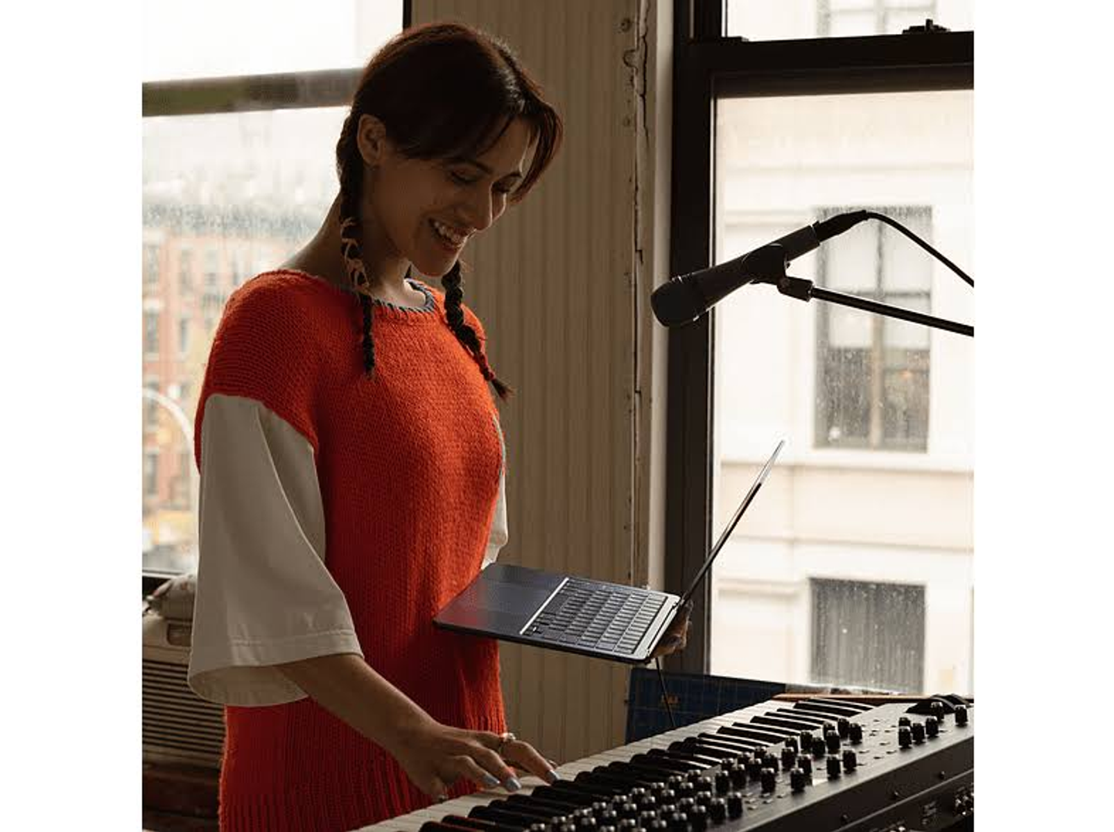 Kobieta gra na klawiaturze, trzyma laptopa. Mikrofon w tle. Kobieta ma na sobie pomarańczowy sweter.